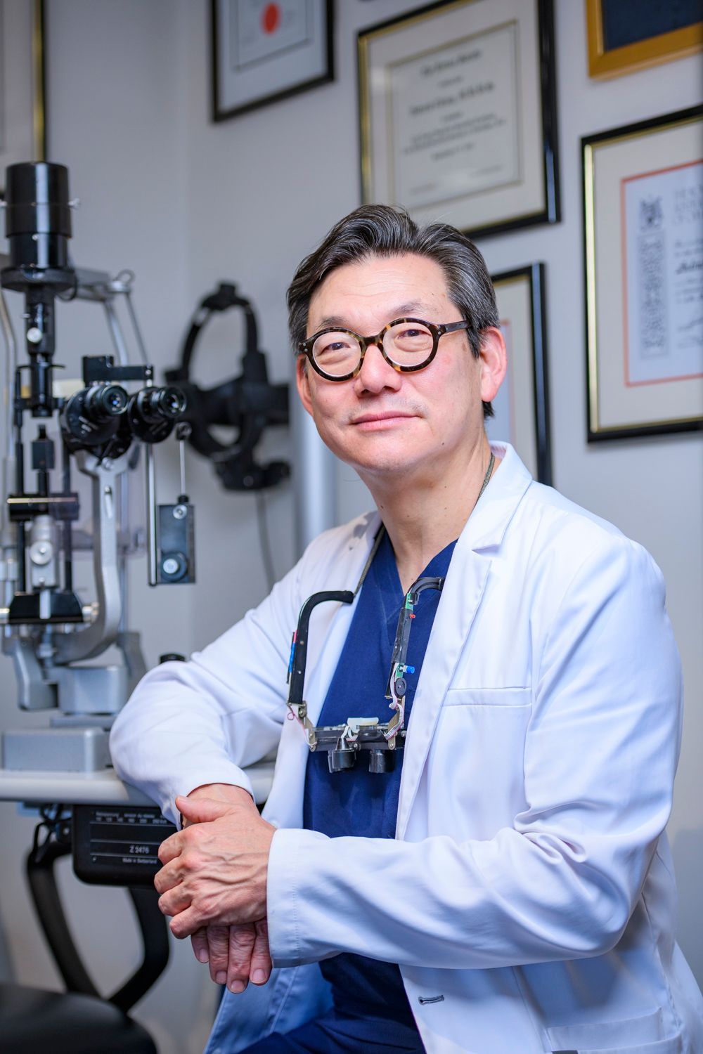 Doctor in glasses and white coat, leaning on eye exam equipment, smiling in an office.