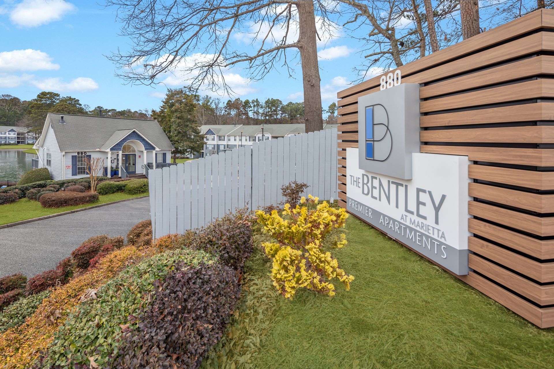 Sign for The Bentley Apartments, white fence, houses, landscaping.