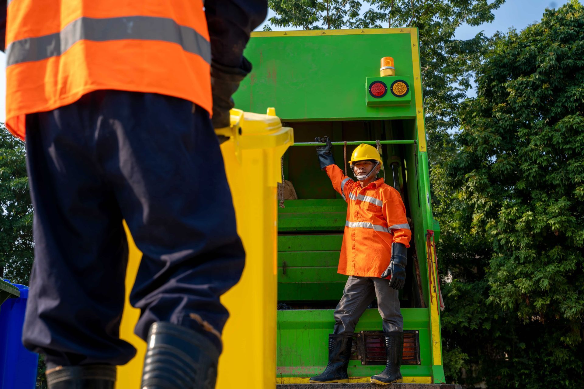 Two sanitation workers emptying a yellow bin into a green garbage truck. One worker directs with his hand up.