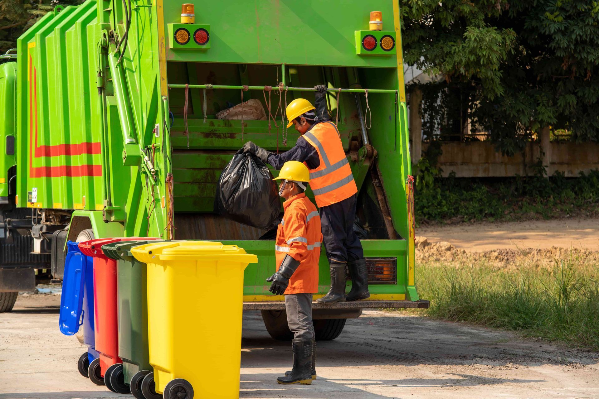 Garbage truck with workers loading a black trash bag. Yellow, blue, and green recycling bins sit in front.