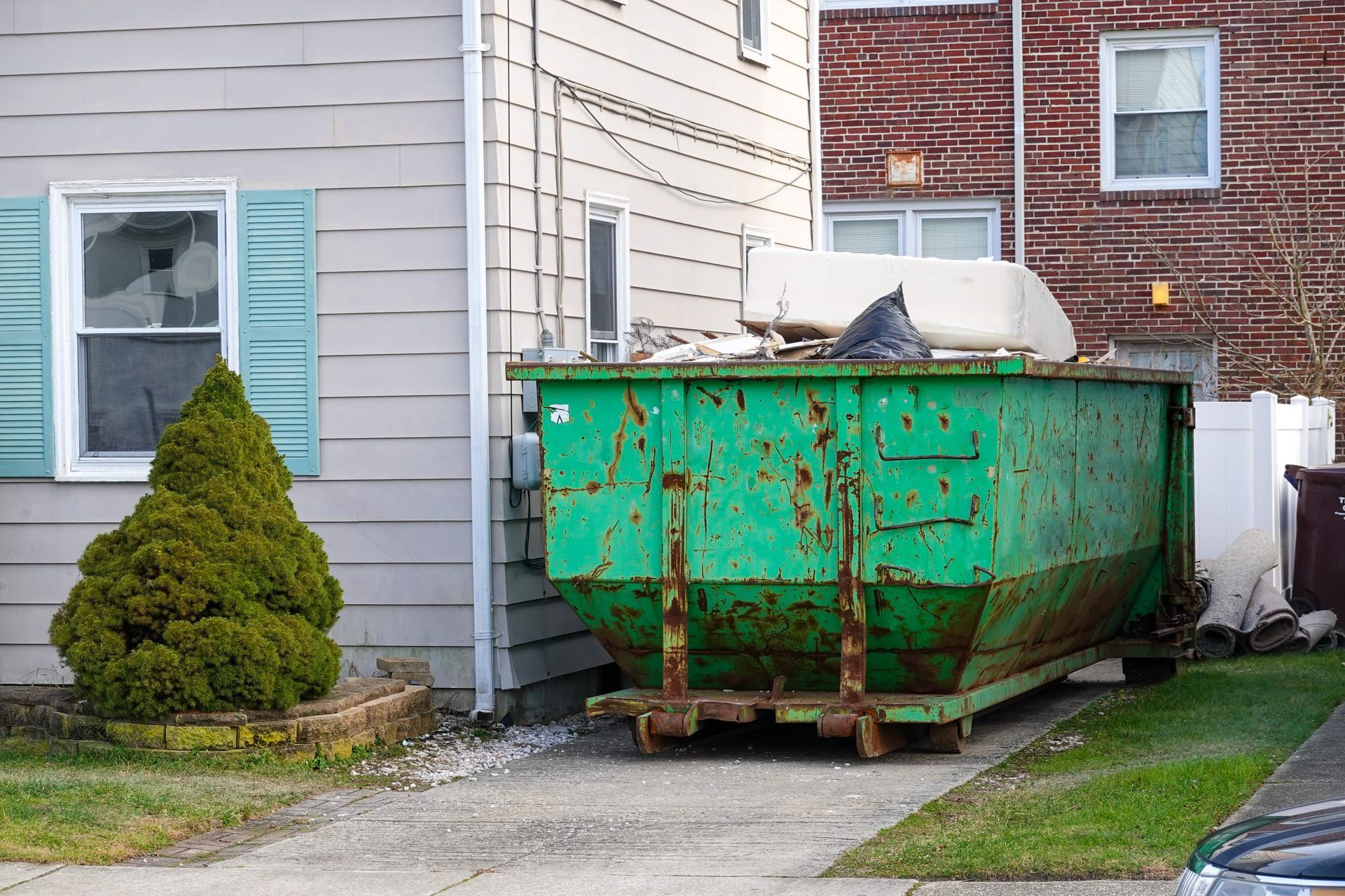 Green dumpster in a driveway next to a light gray house.