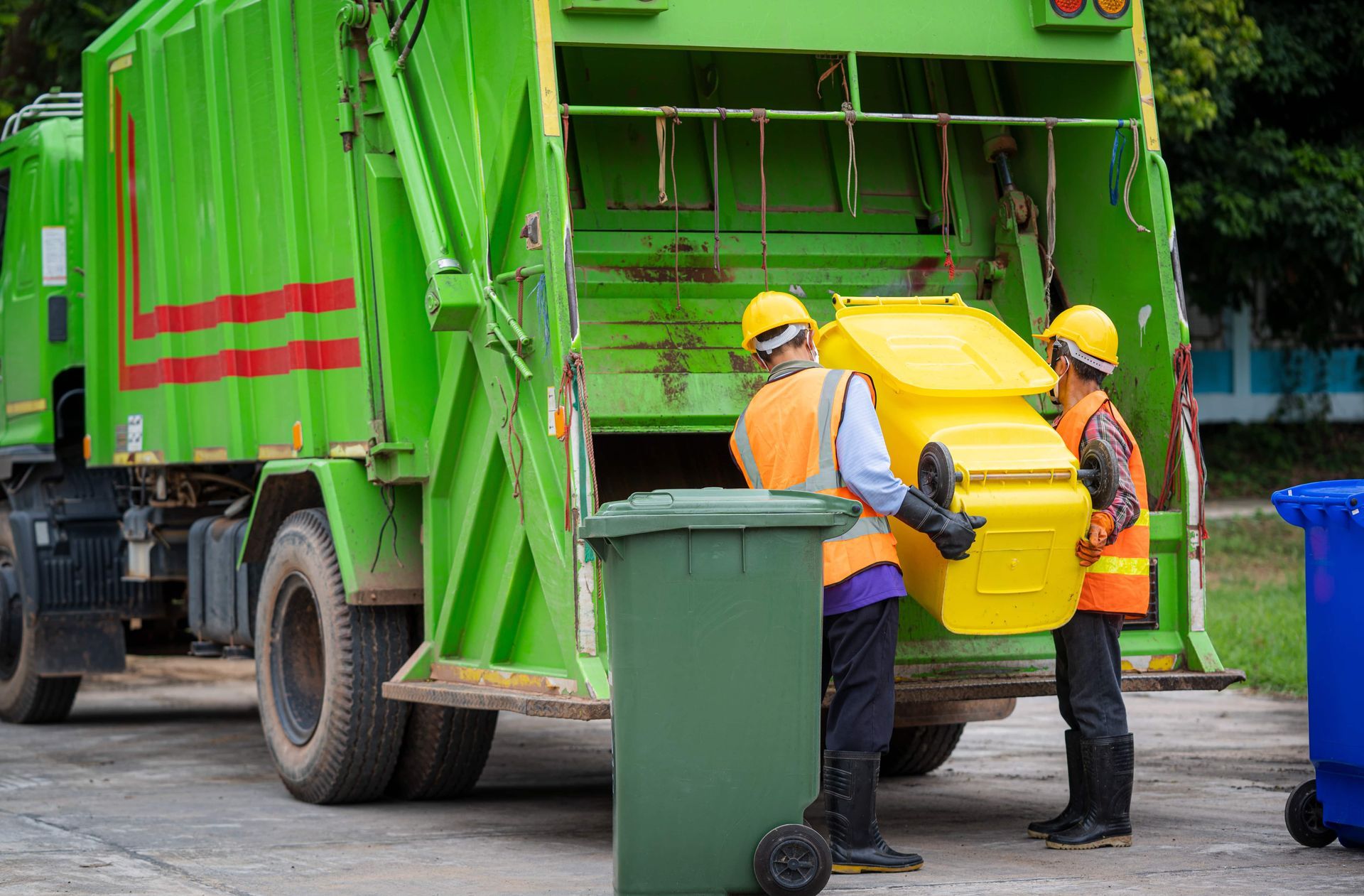 Two sanitation workers loading a yellow recycling bin into a green garbage truck.