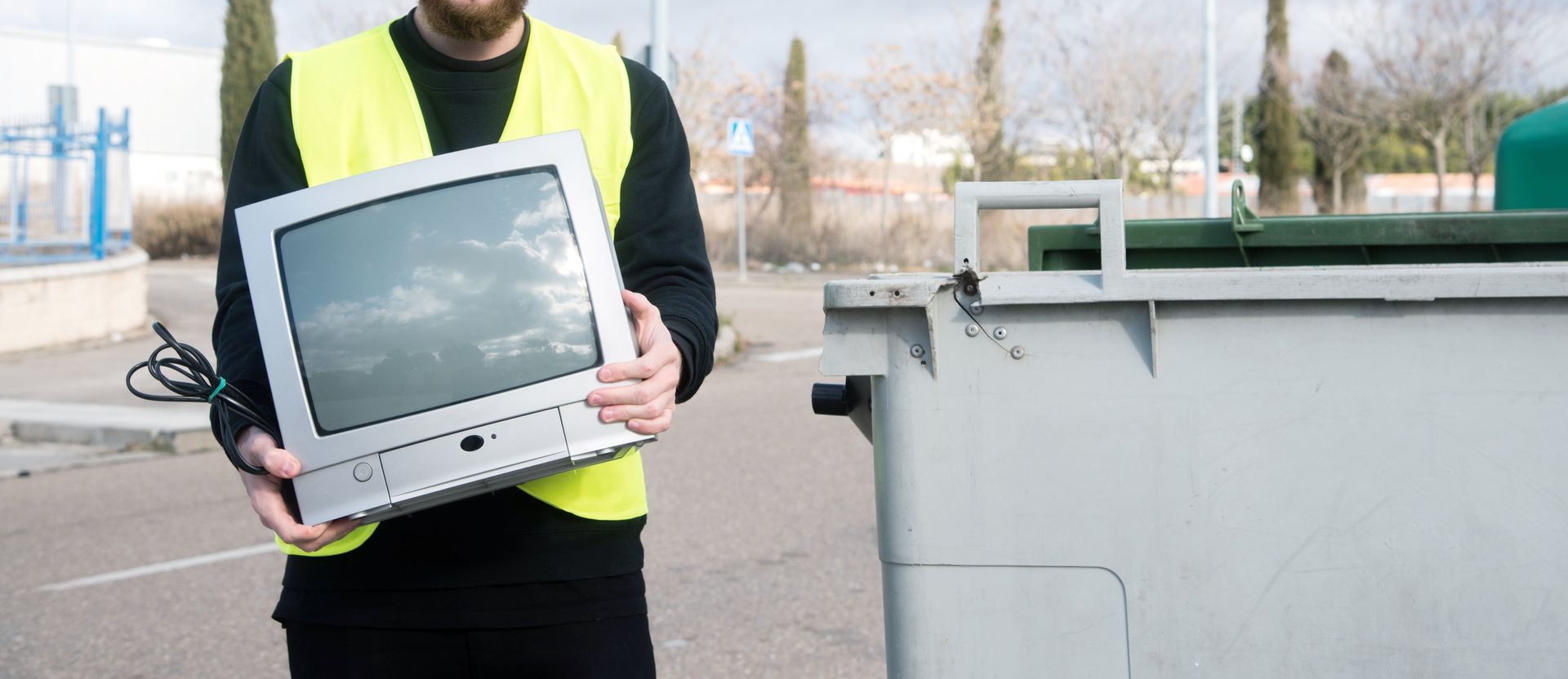 Person in a yellow vest holding an old television near a large gray recycling bin.