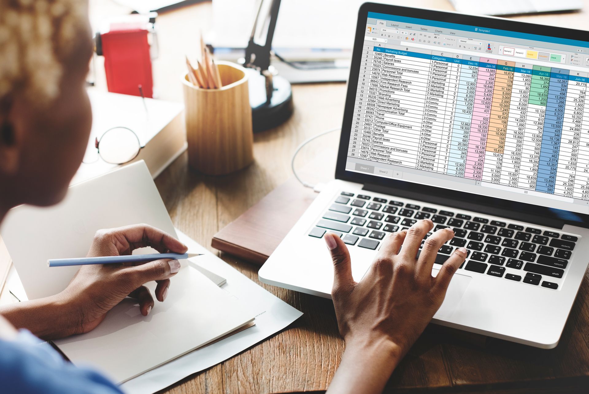 Person working on a laptop with spreadsheet, writing in a notebook, pencils in a cup, on a wooden desk.