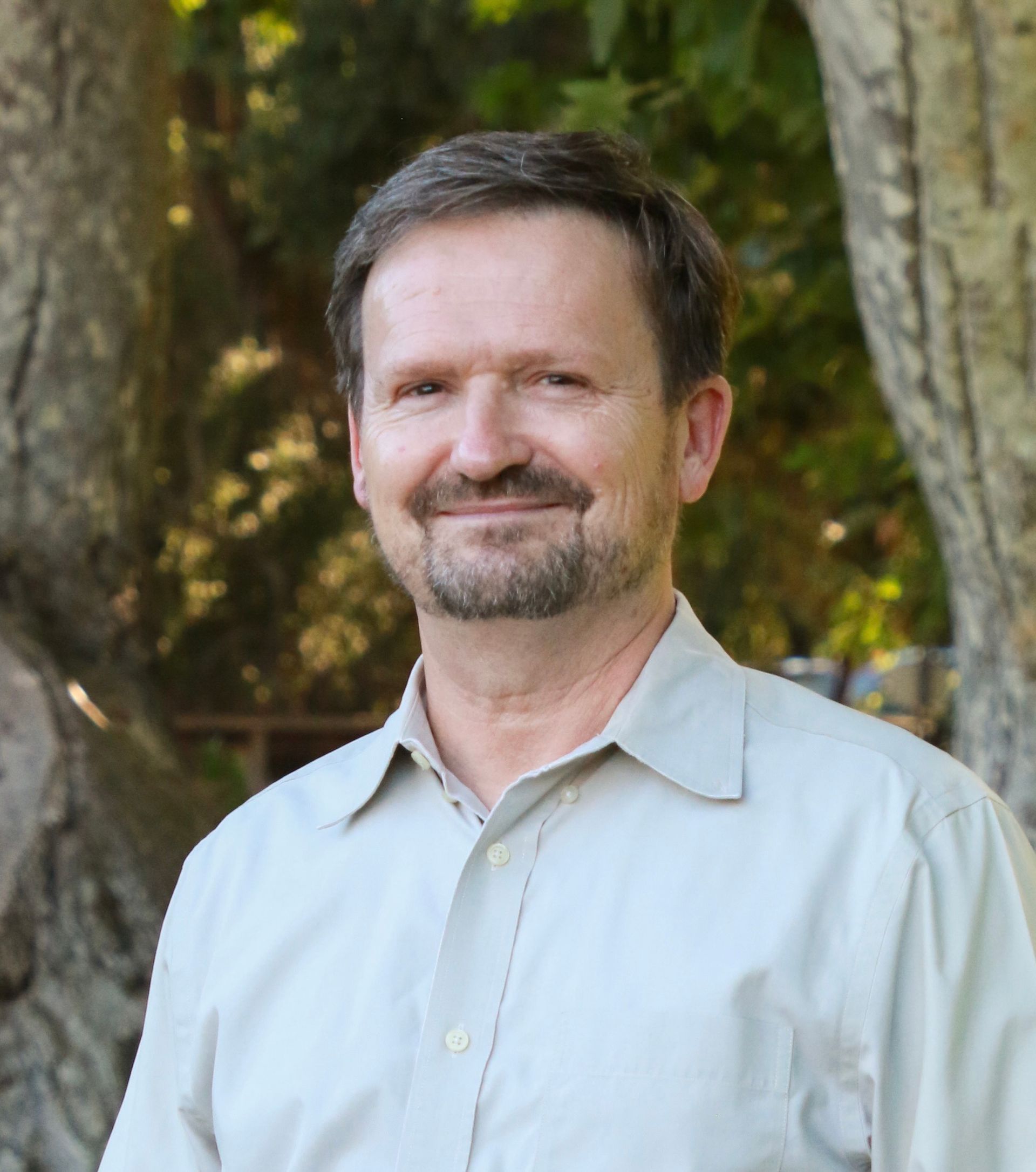 Man with a goatee smiling, wearing a light-colored button-down shirt outdoors near trees.