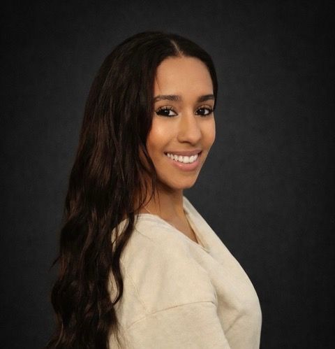 Woman with long wavy brown hair, smiling, looking towards the camera, wearing a cream sweater, dark background.