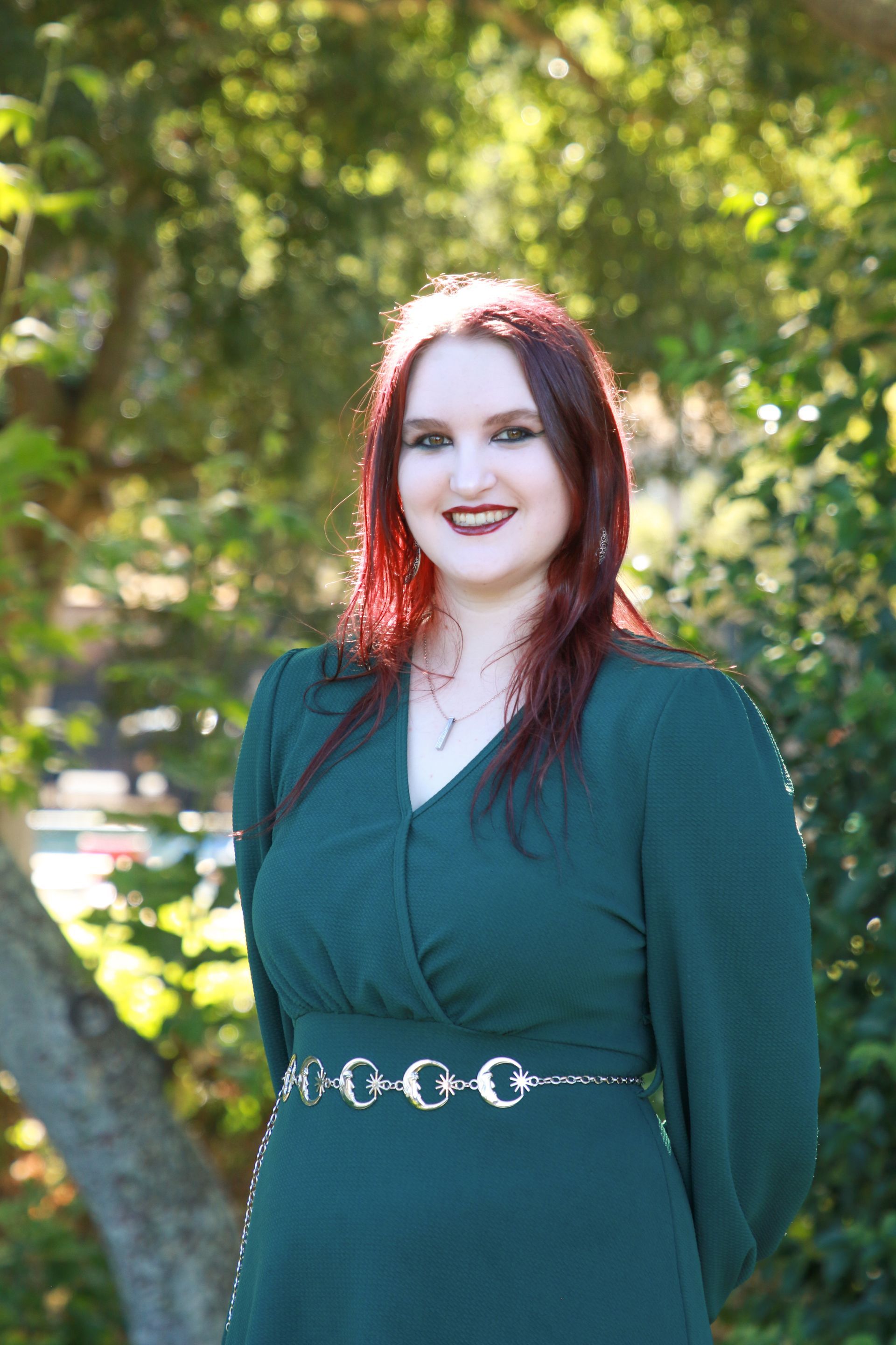 Woman with red-tinted hair wearing a green dress and silver belt, smiling in a garden.