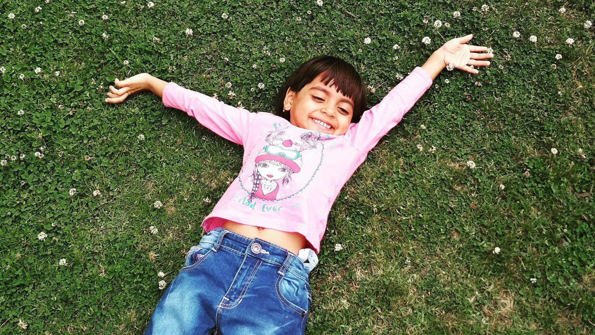 A little girl is laying on the grass with her arms outstretched.