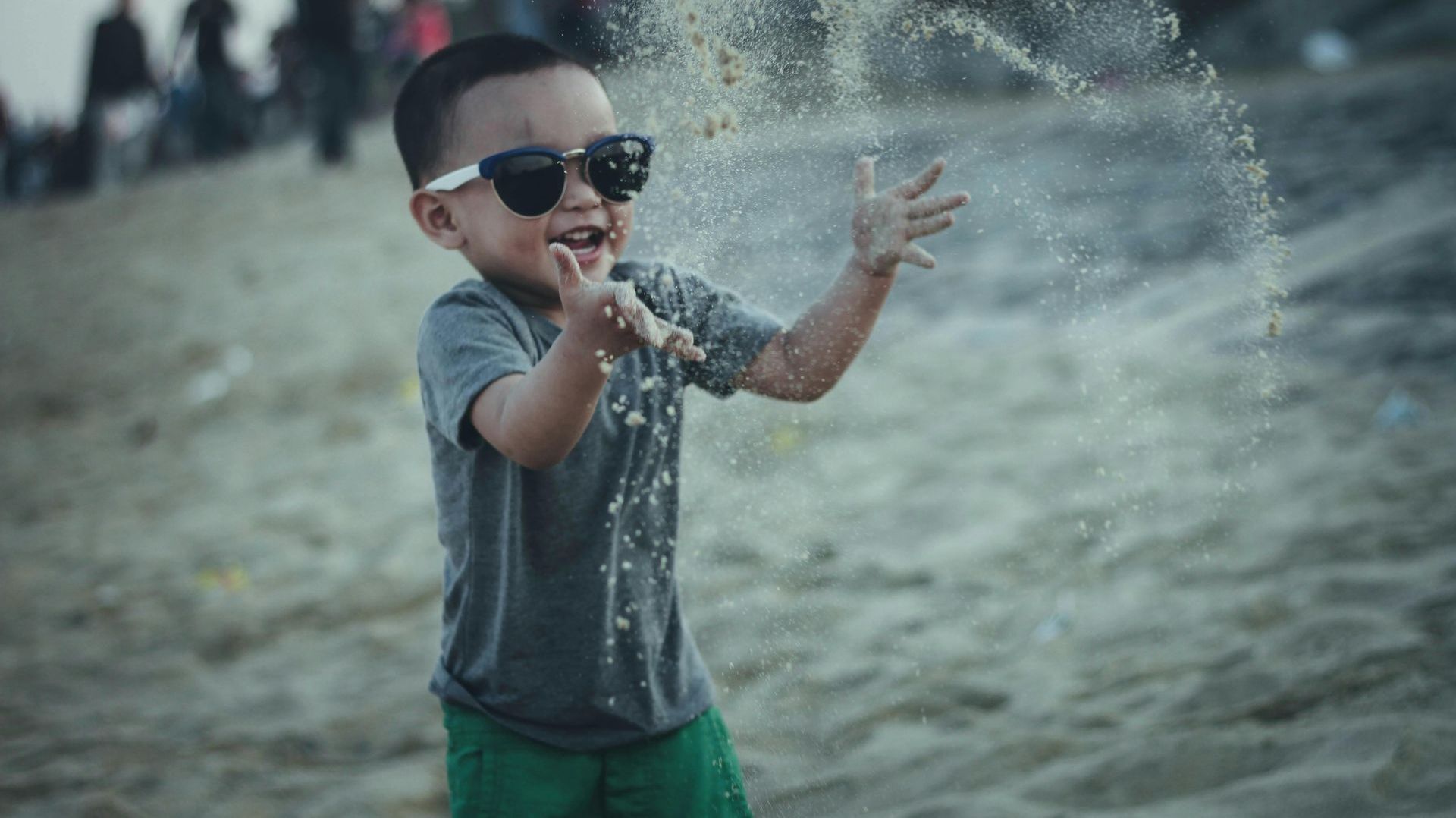 A young boy wearing sunglasses is playing with sand on the beach.