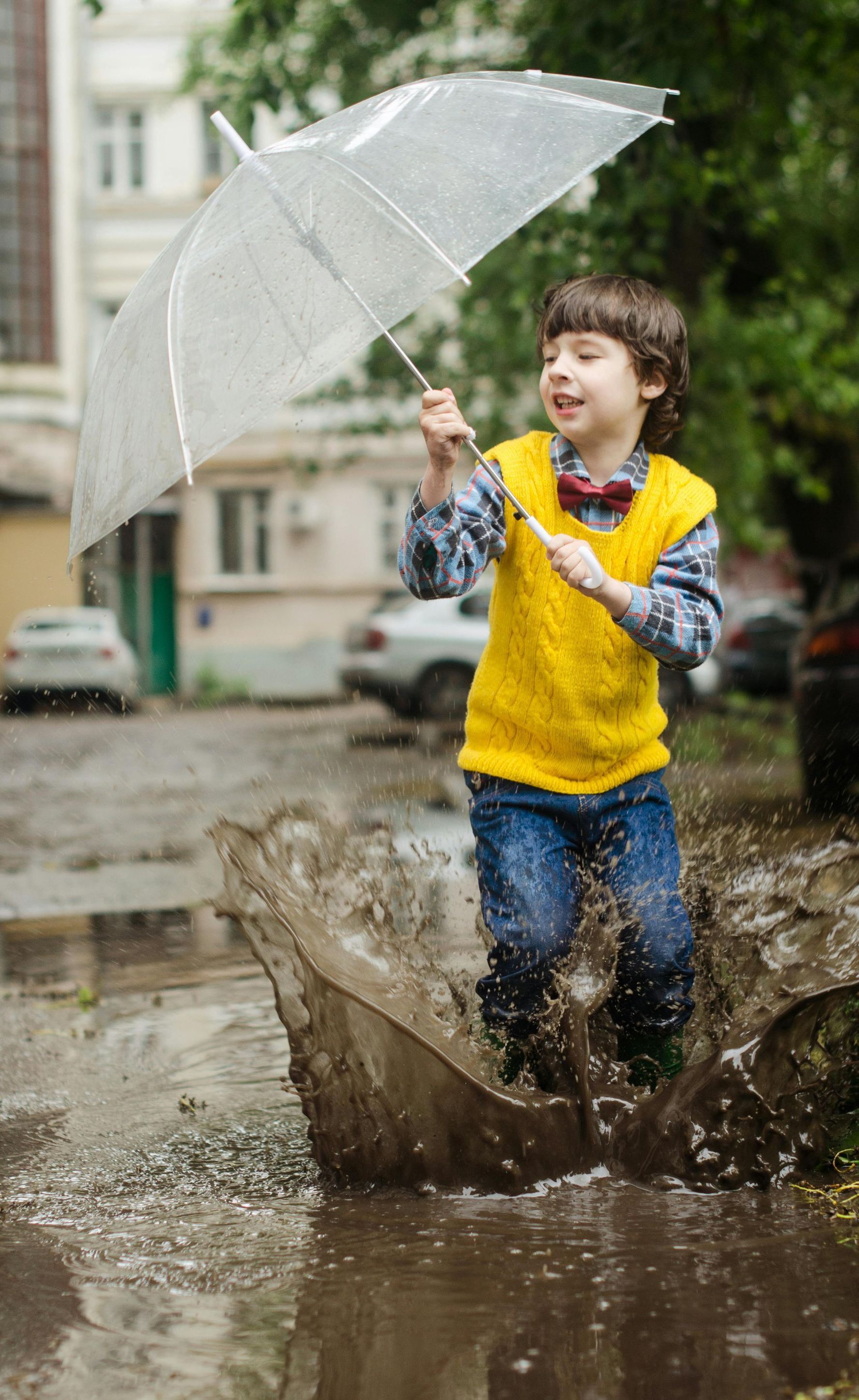 A young boy is holding an umbrella and jumping into a puddle.