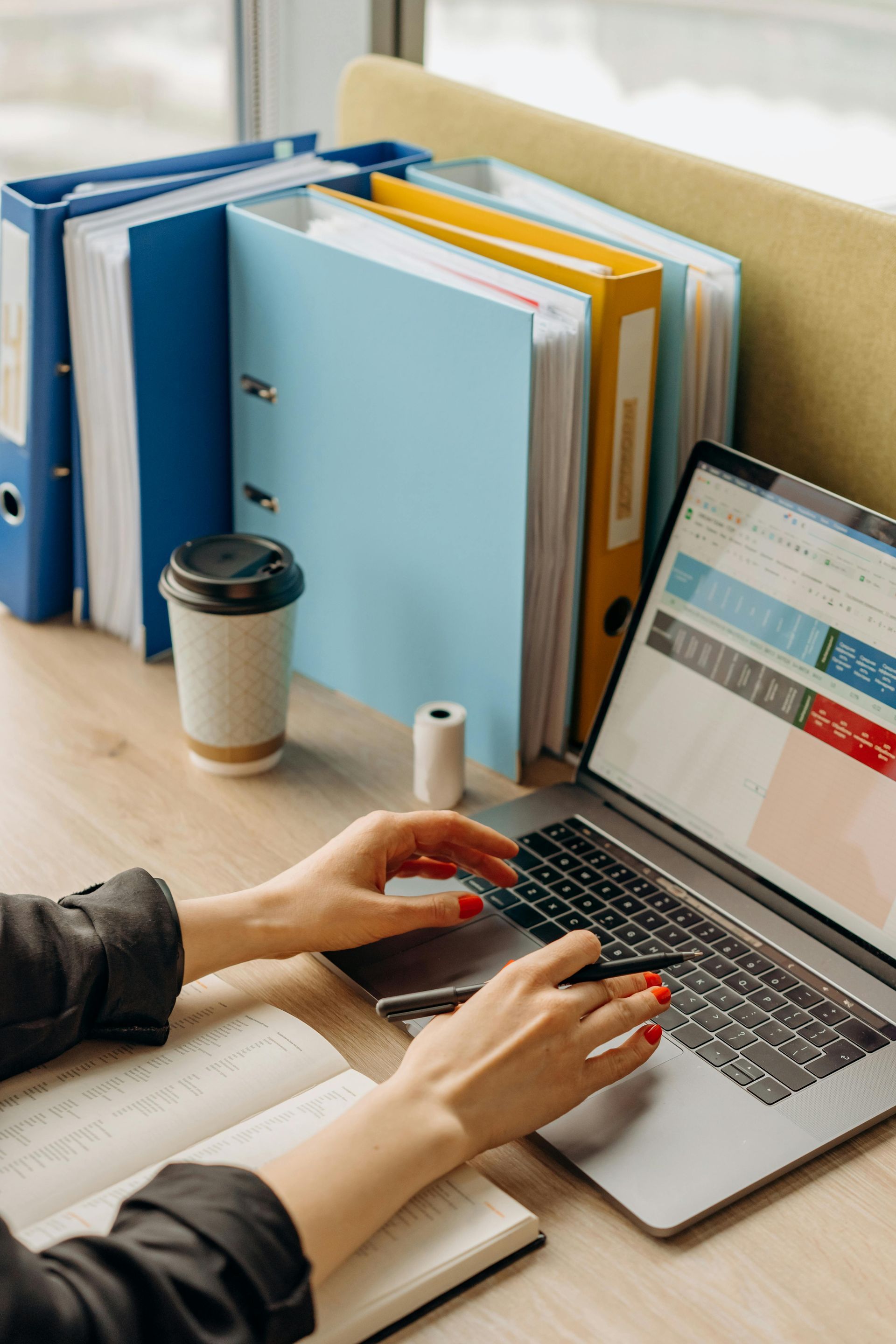 Hands typing on laptop at desk with folders, coffee cup, and notebook.