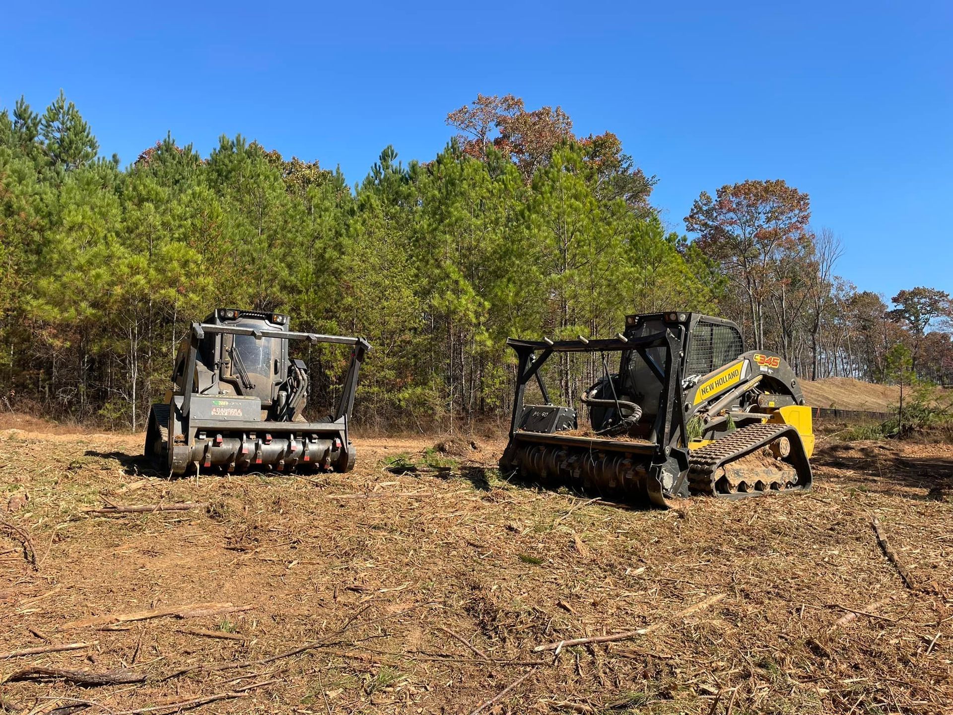 Two forestry mulchers in a cleared field, with forest in the background on a sunny day.