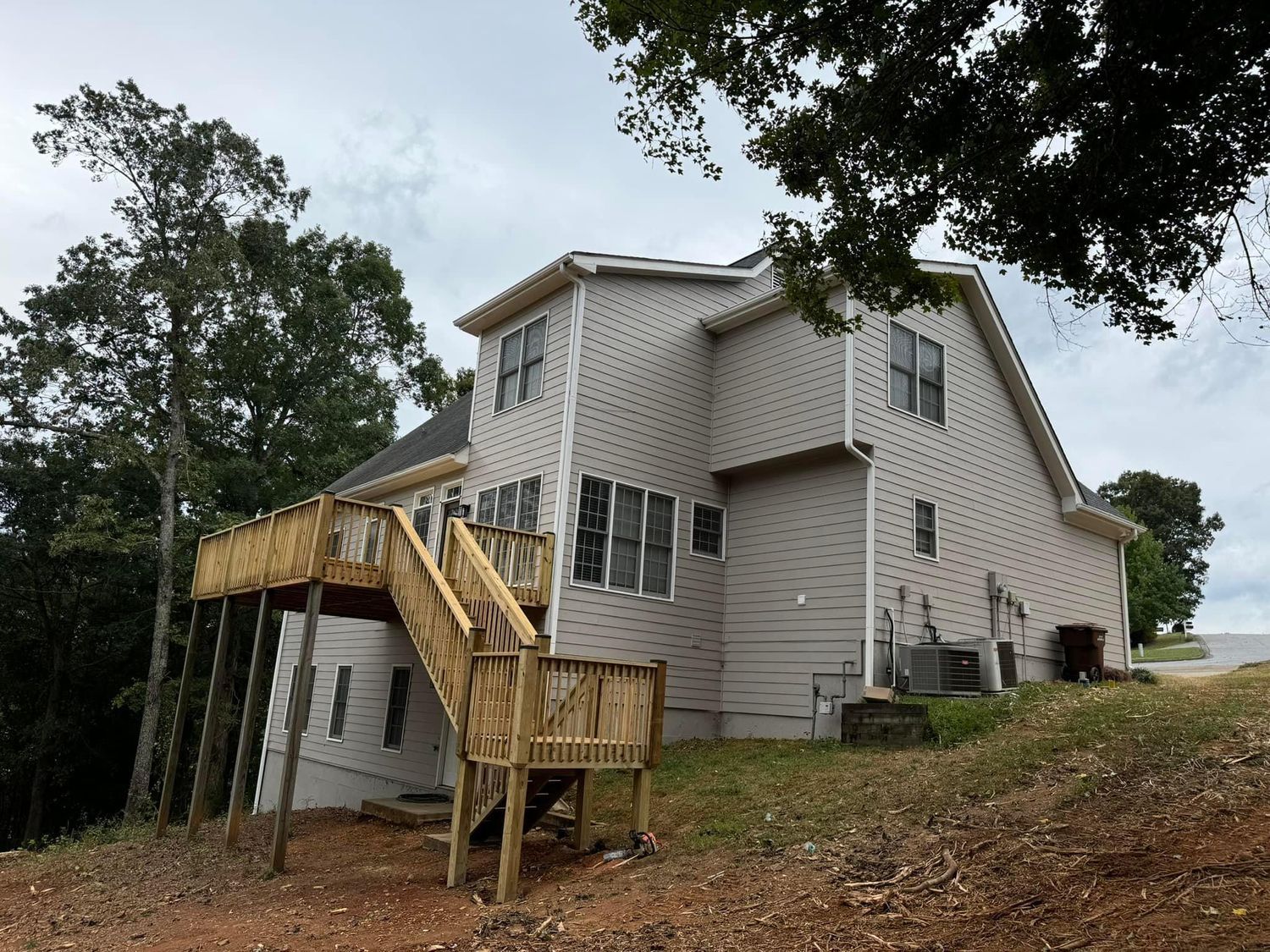 Two-story house with wooden deck and stairs on a hillside under a cloudy sky.