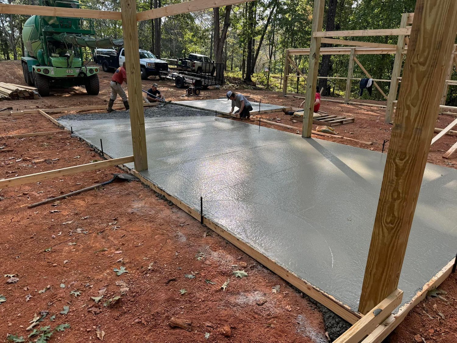 Construction site with workers pouring fresh concrete floor. Wooden frame, trees, mixer, and trucks visible.