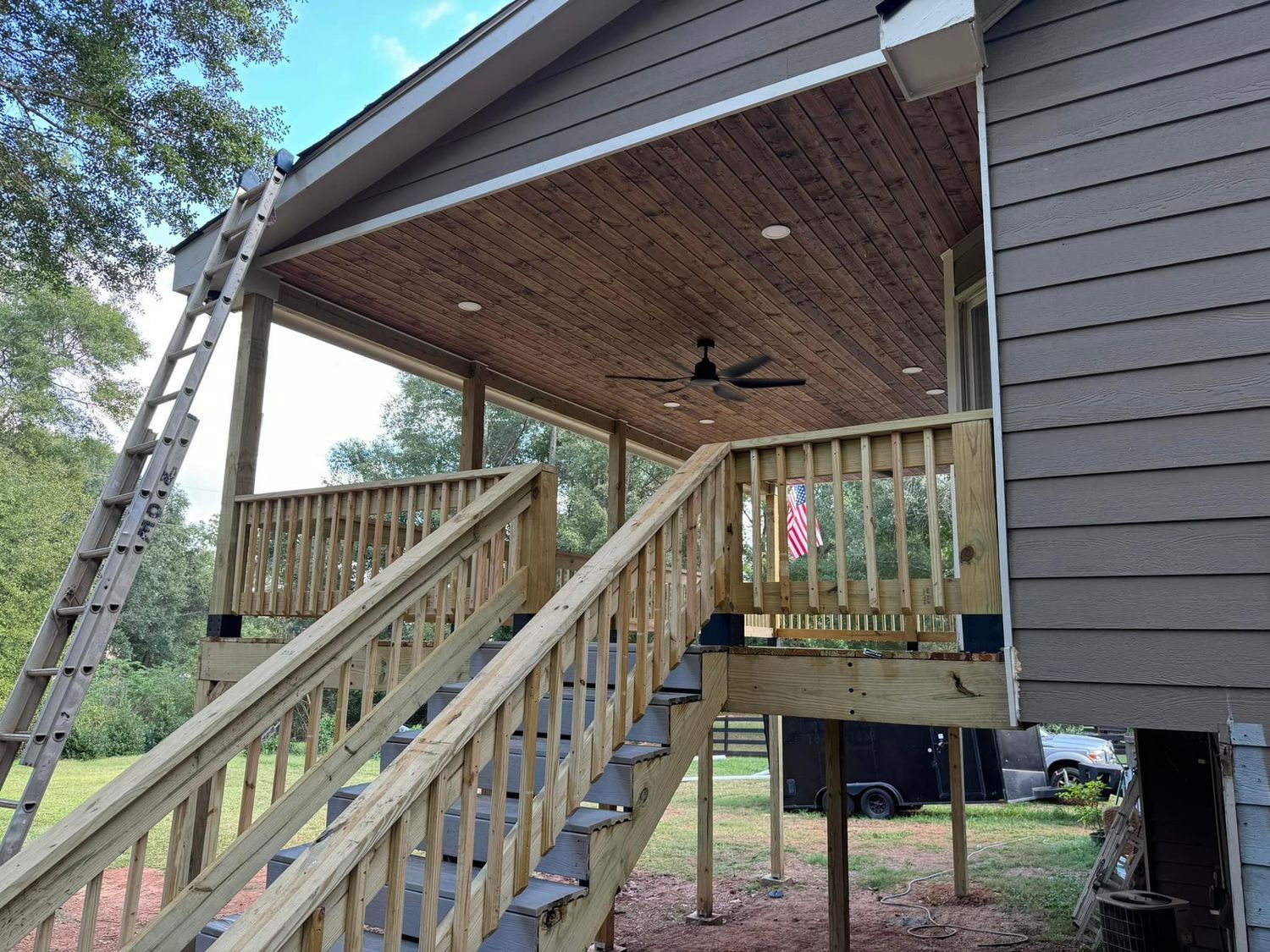 Wooden porch with stairs, railing, and a brown ceiling; ladder leans against the roof.
