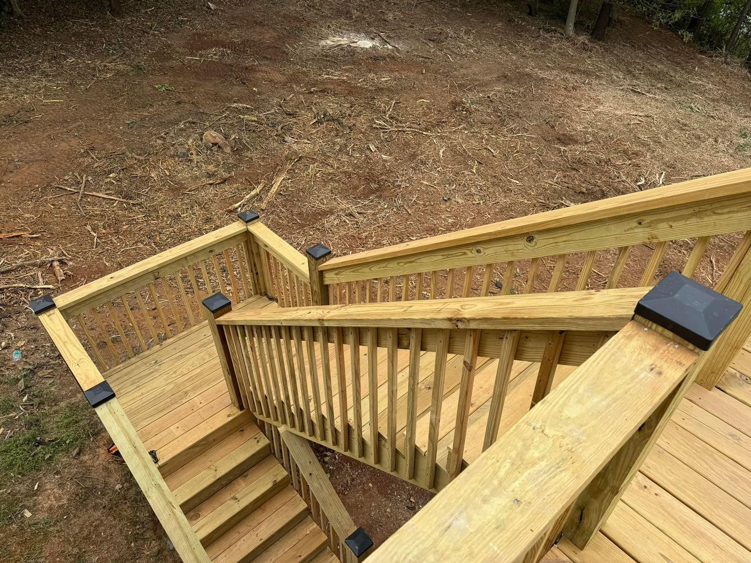 Wooden outdoor staircase with railings, black post caps, surrounded by a sloped, wood-chipped yard.