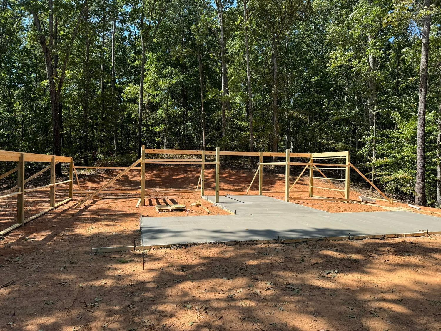 Construction site: concrete pad with wooden frame, set in a wooded area under a blue sky.