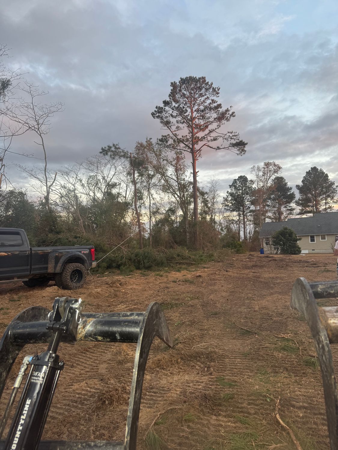 Clearing land: A truck and heavy machinery are on brown soil near a wooded area with a house visible in the distance under a cloudy sky.