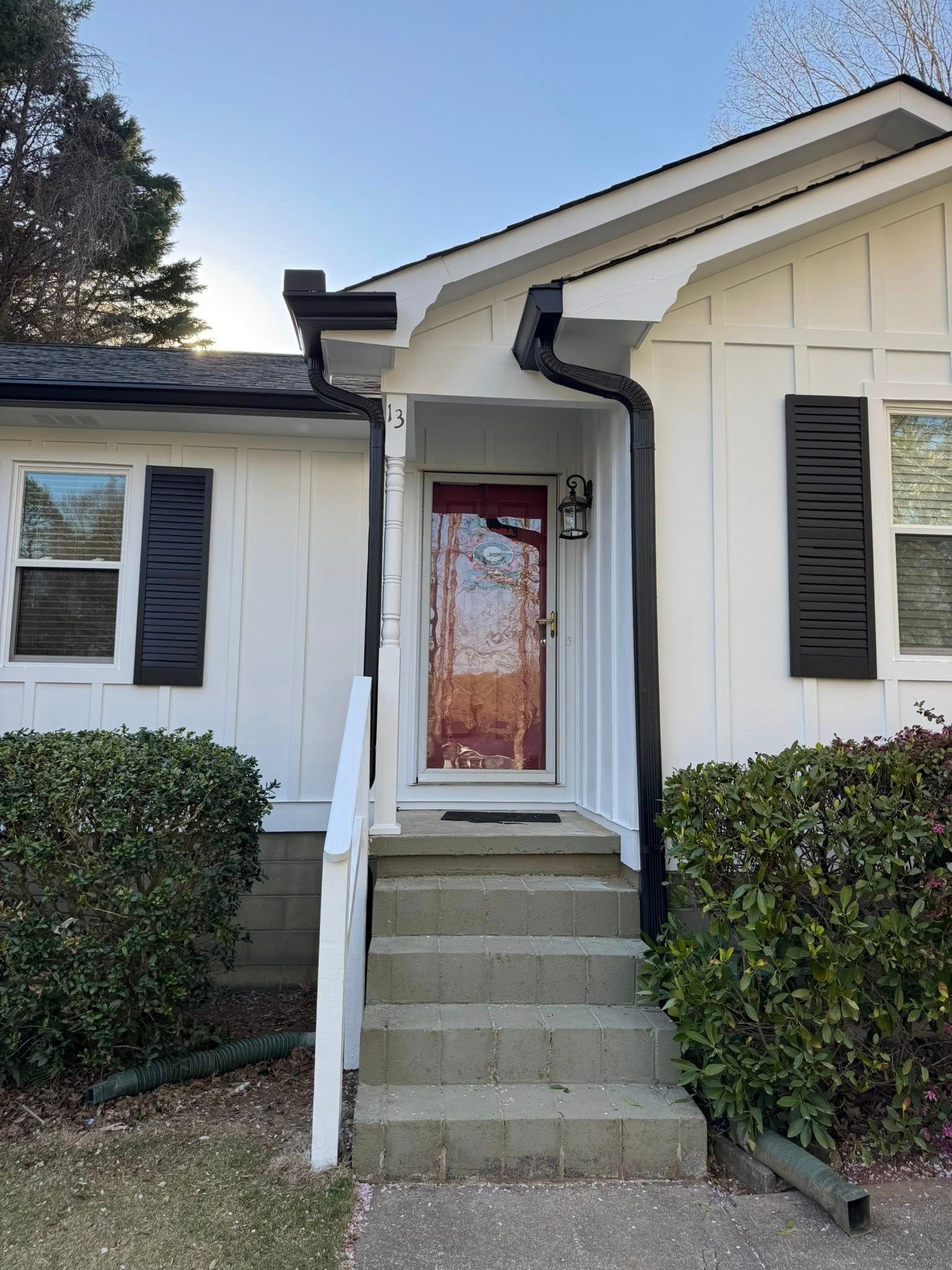White house with black shutters, steps to a red door, bushes on either side.
