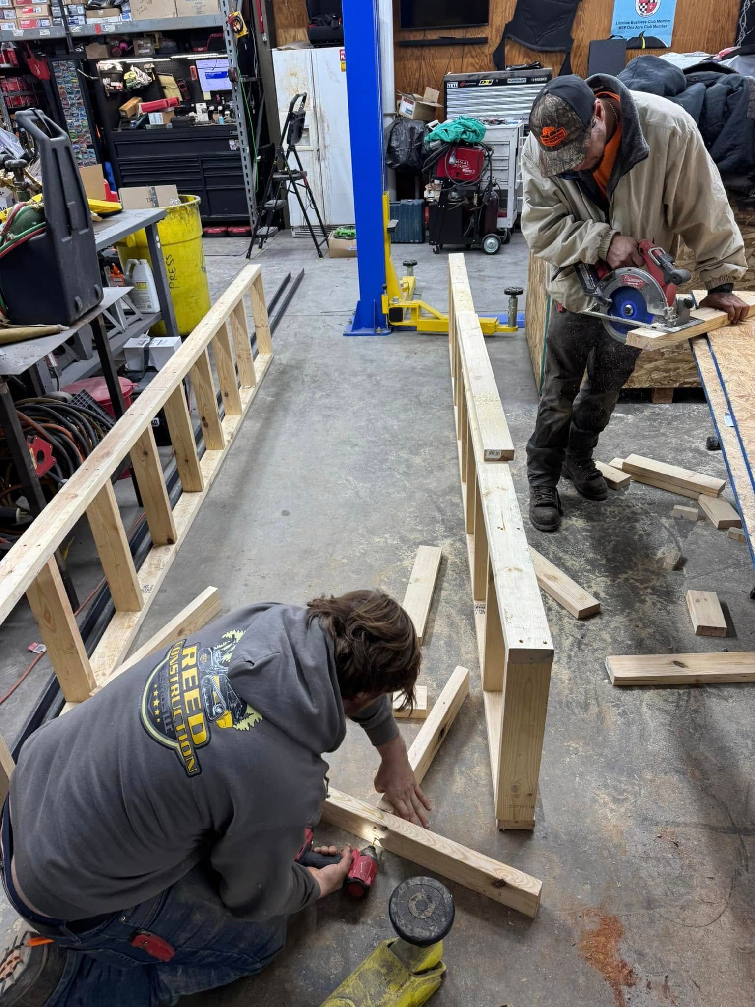 Two people building a wooden structure in a workshop. One cuts wood with a saw while the other uses a drill.