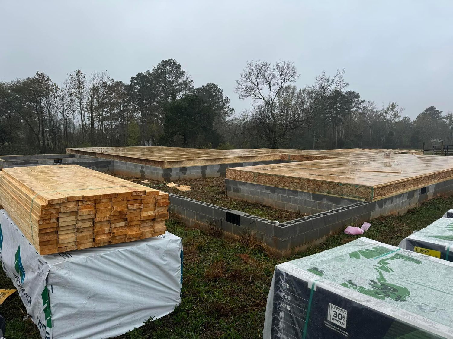Construction site with concrete block foundations and wood flooring in progress, lumber stacked nearby on a cloudy day.