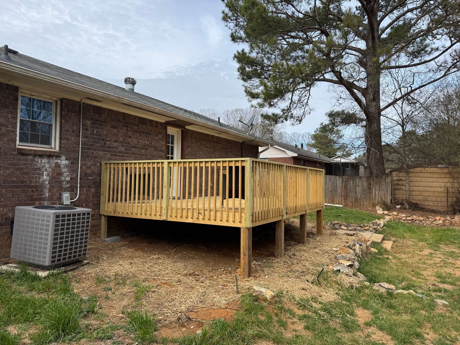 Wooden deck attached to a brick house, gravel ground, tree, and air conditioning unit.