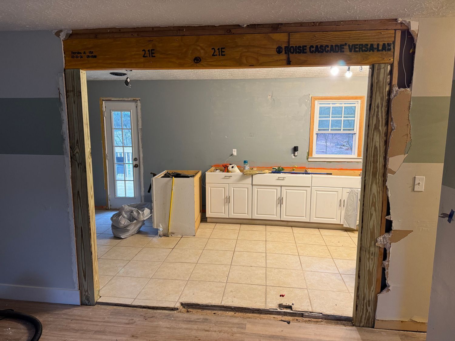 Renovated kitchen doorway; wood beam and framing. Kitchen visible with white cabinets and a window.