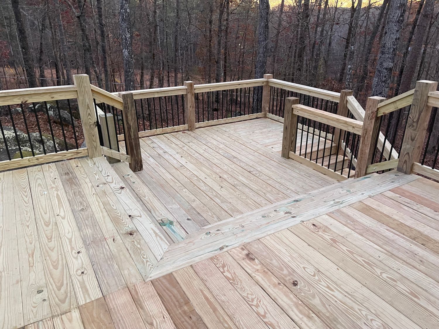 Wooden deck with railings, steps, and black spindles in a wooded area.