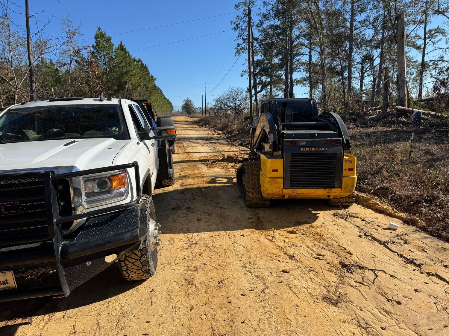 A white truck and yellow skid steer on a dirt road in a wooded area on a sunny day.