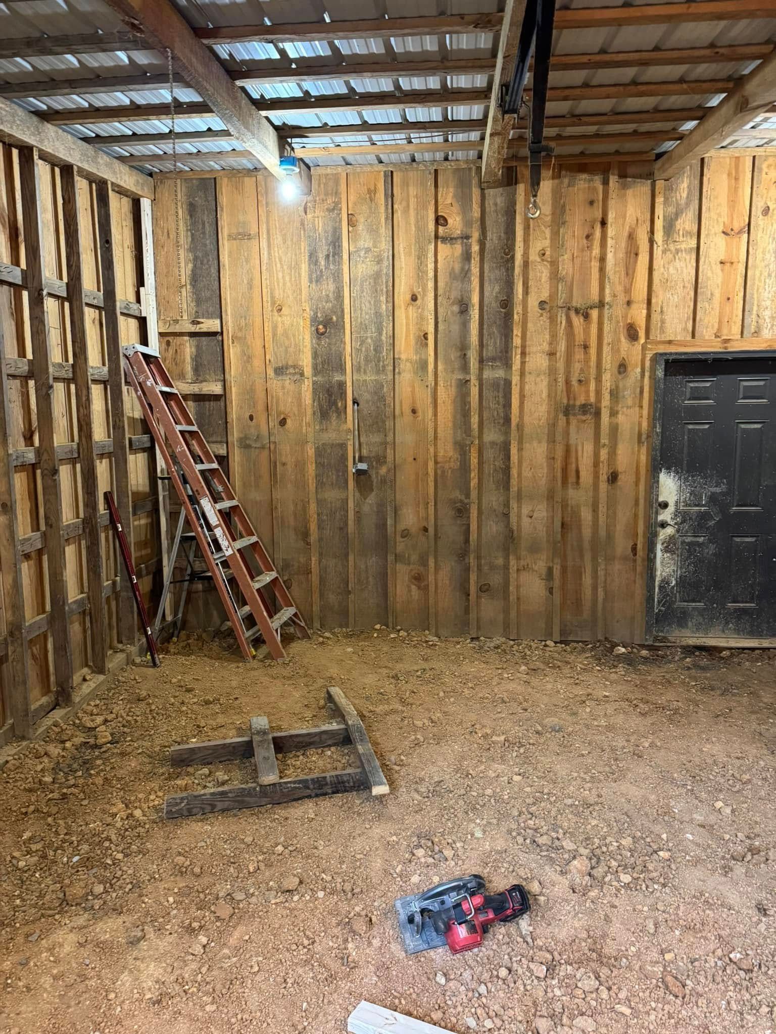 Interior of a barn with wood paneling walls and dirt floor. A ladder leans against the wall, tools on the floor.