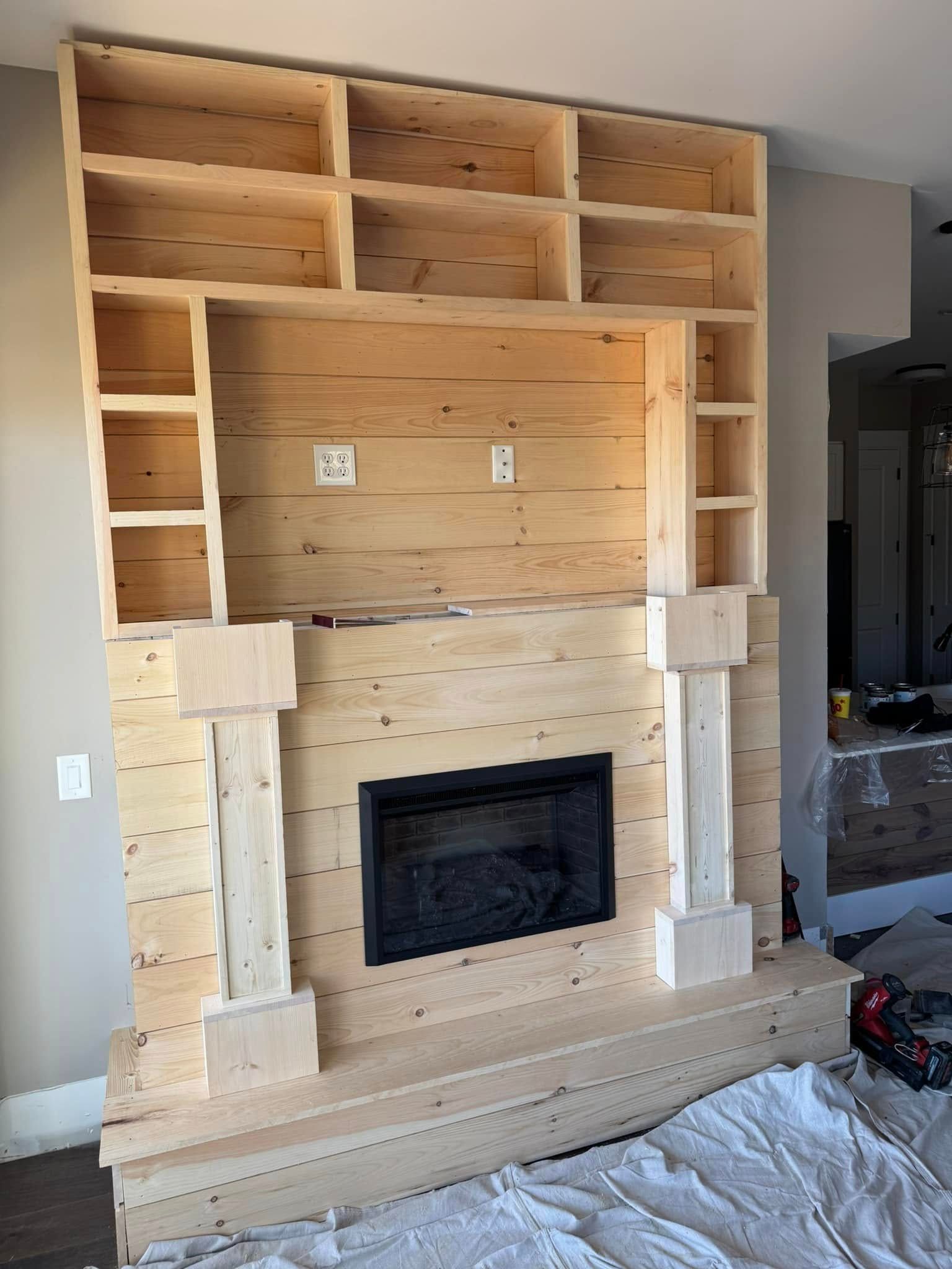 Unfinished wooden fireplace with built-in bookshelves, a dark fireplace insert, and unfinished wood on a construction site.