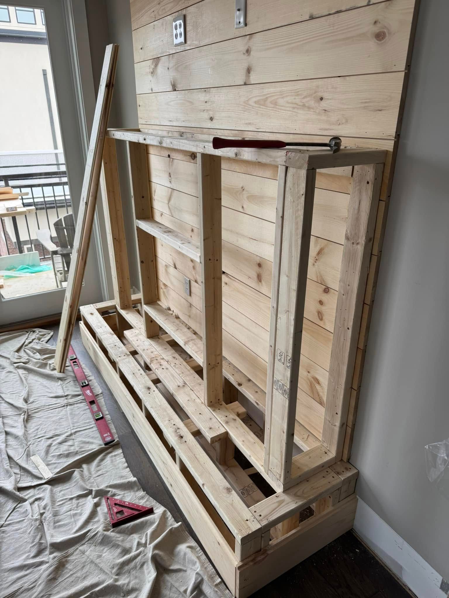 Wooden framework of a built-in entertainment center, under construction against a wood-paneled wall, near a window.