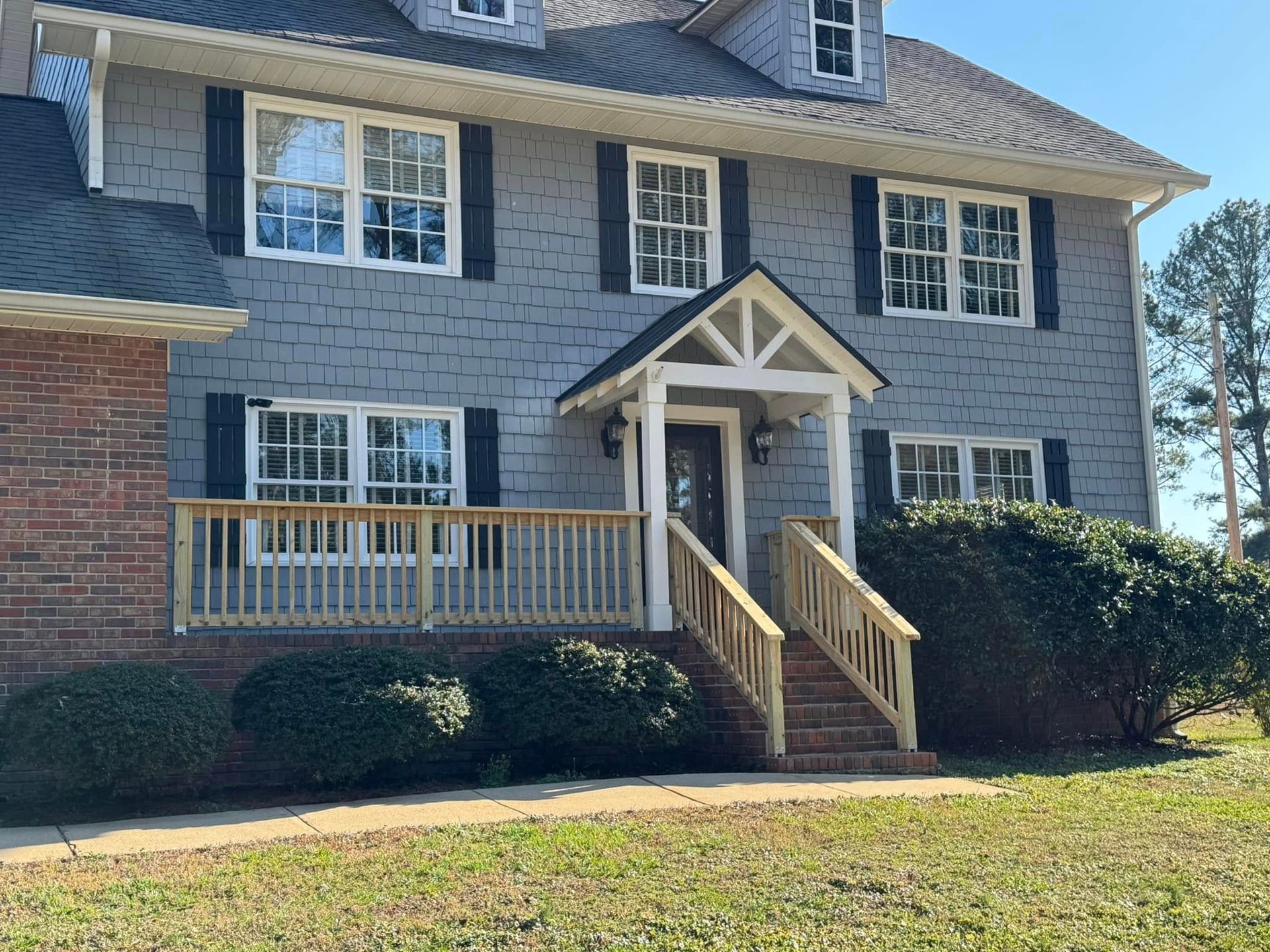 Two-story house with gray siding, wooden porch, and bushes in front. Blue sky and brick on the left.