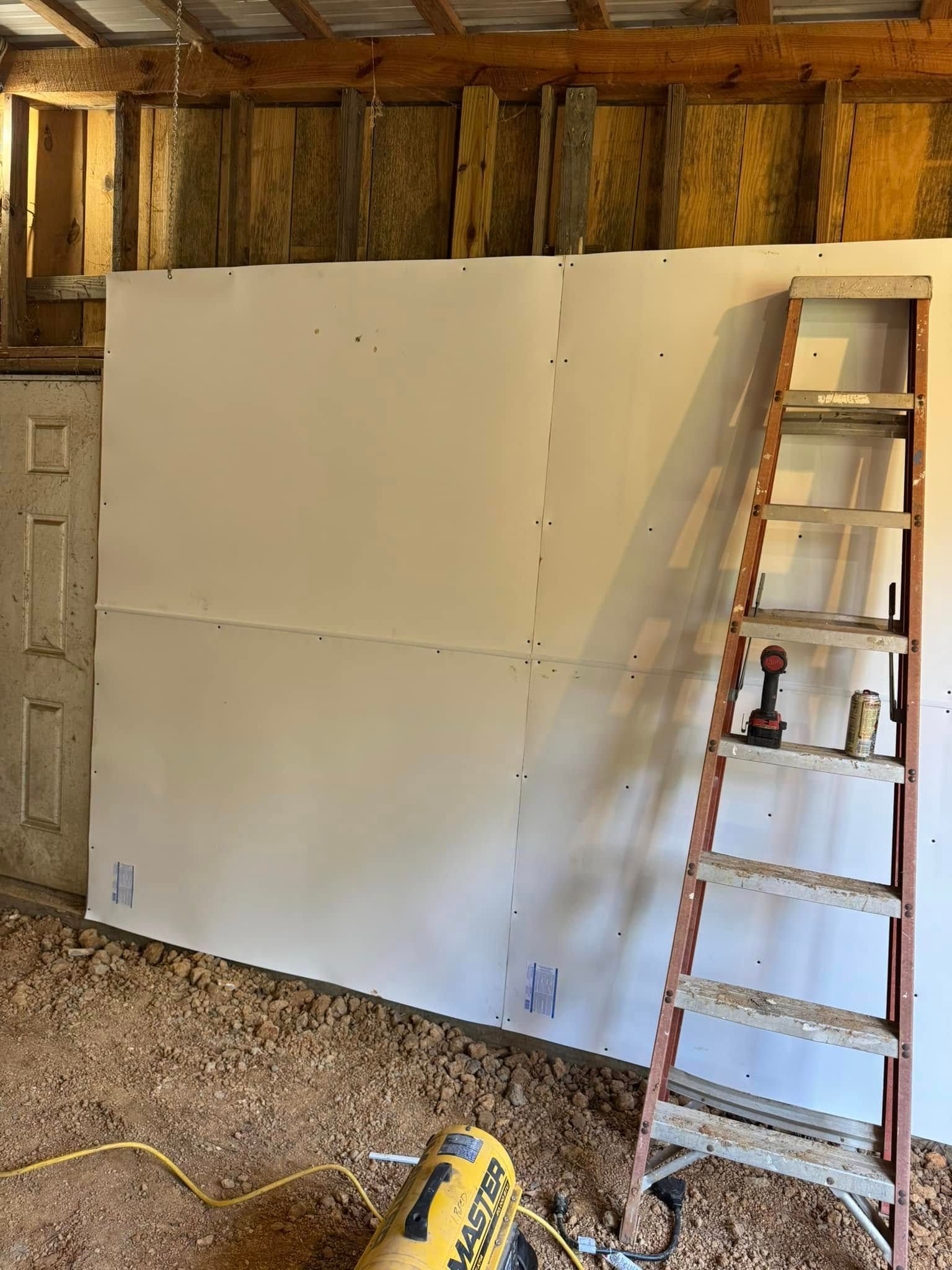 Drywall installation on a wall in a building with a ladder, insulation, and a heater on the floor.