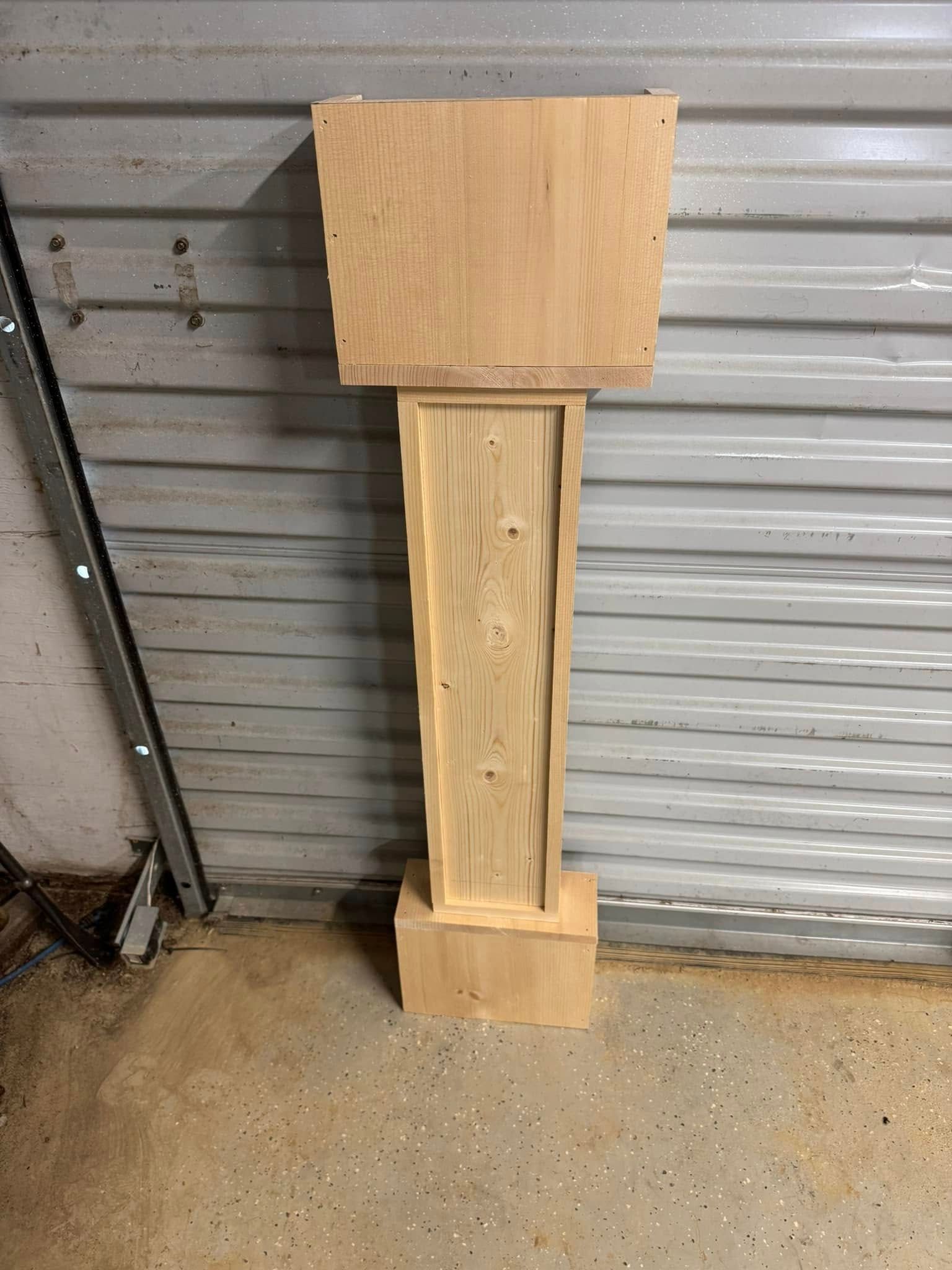 Unfinished wooden grandfather clock base with a rectangular top and base, set against a corrugated metal wall.