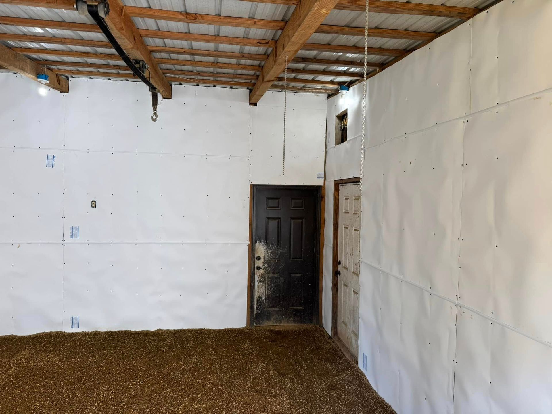 Interior of a barn with white paneled walls, a dark door, and a wooden ceiling. The floor is covered in brown material.