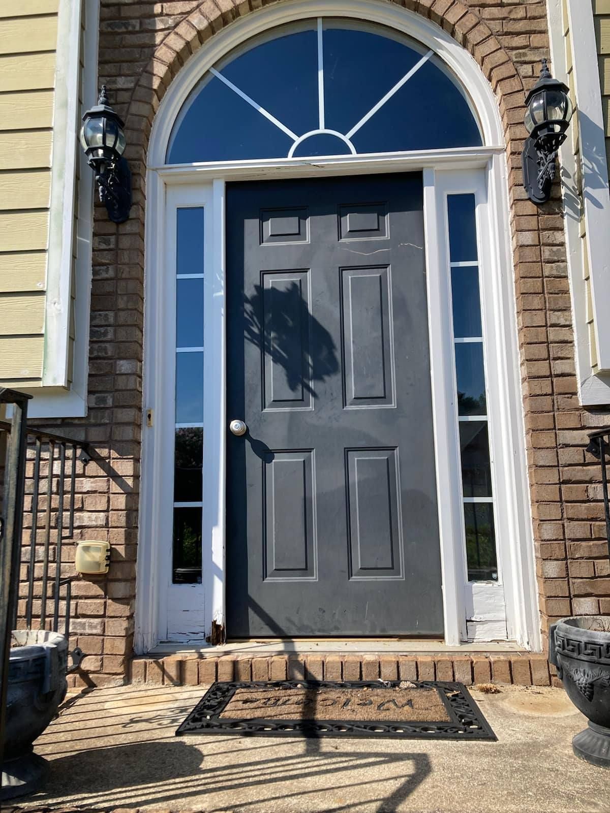 Front door painted dark gray with arched window above and sidelights. Brick exterior, black sconces, welcome mat.