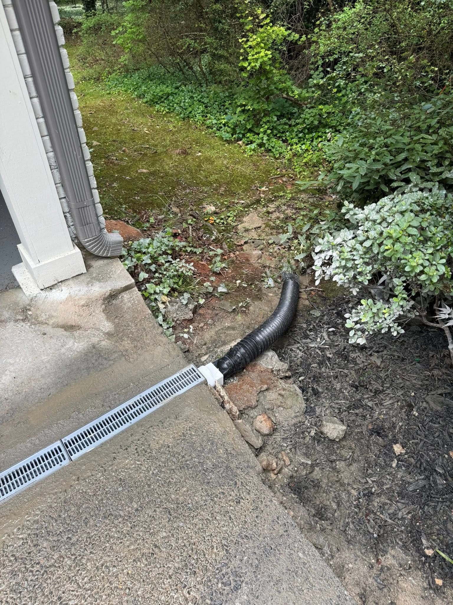 A downspout directs water to a black curved pipe on a concrete step, near a bush and grassy area.