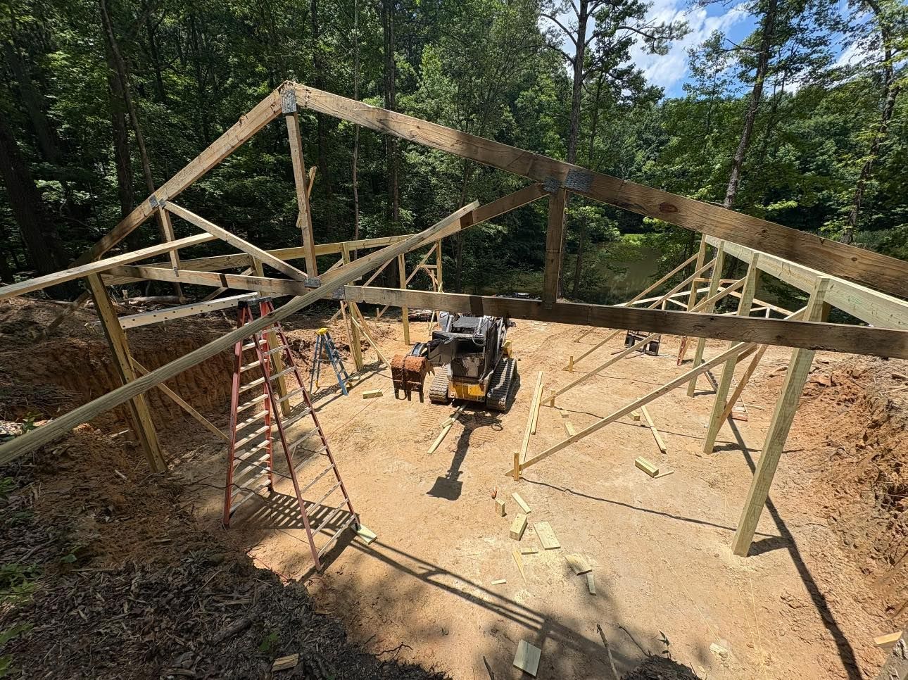 Construction of a wooden building frame in a wooded area; a small excavator is in the dirt foundation.