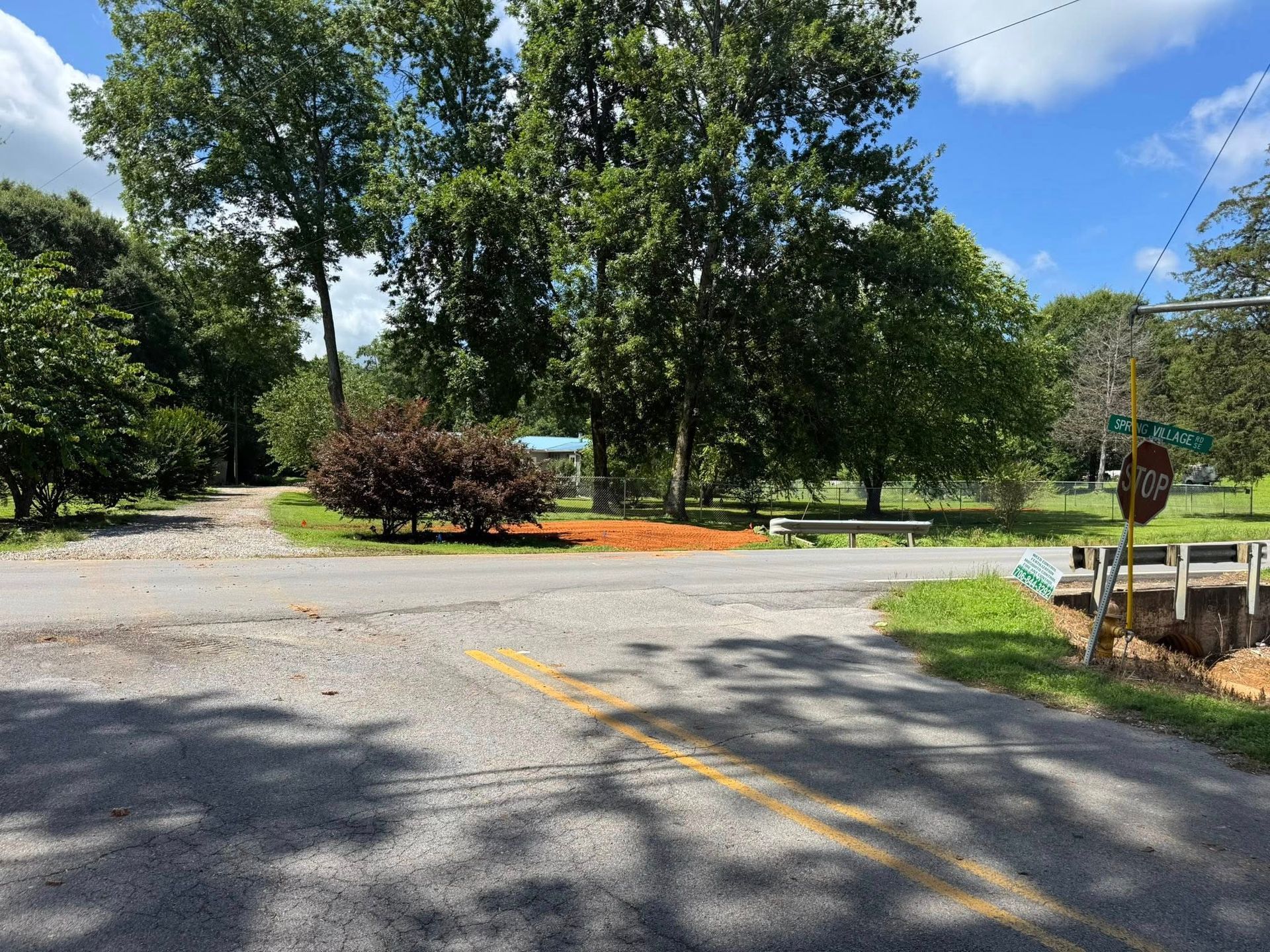 Gravel road intersection with trees, bushes, and a green sign against a blue sky.