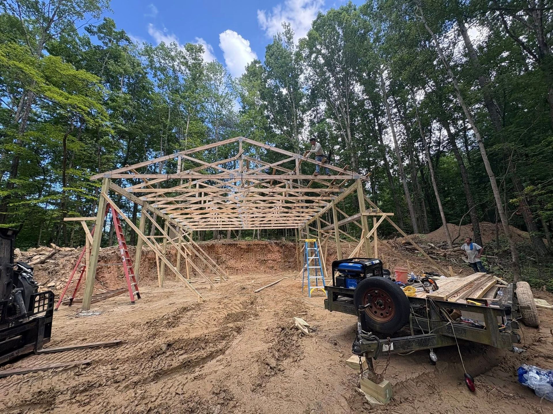 Wooden structure of a barn being built in a forest, with construction materials and equipment nearby.
