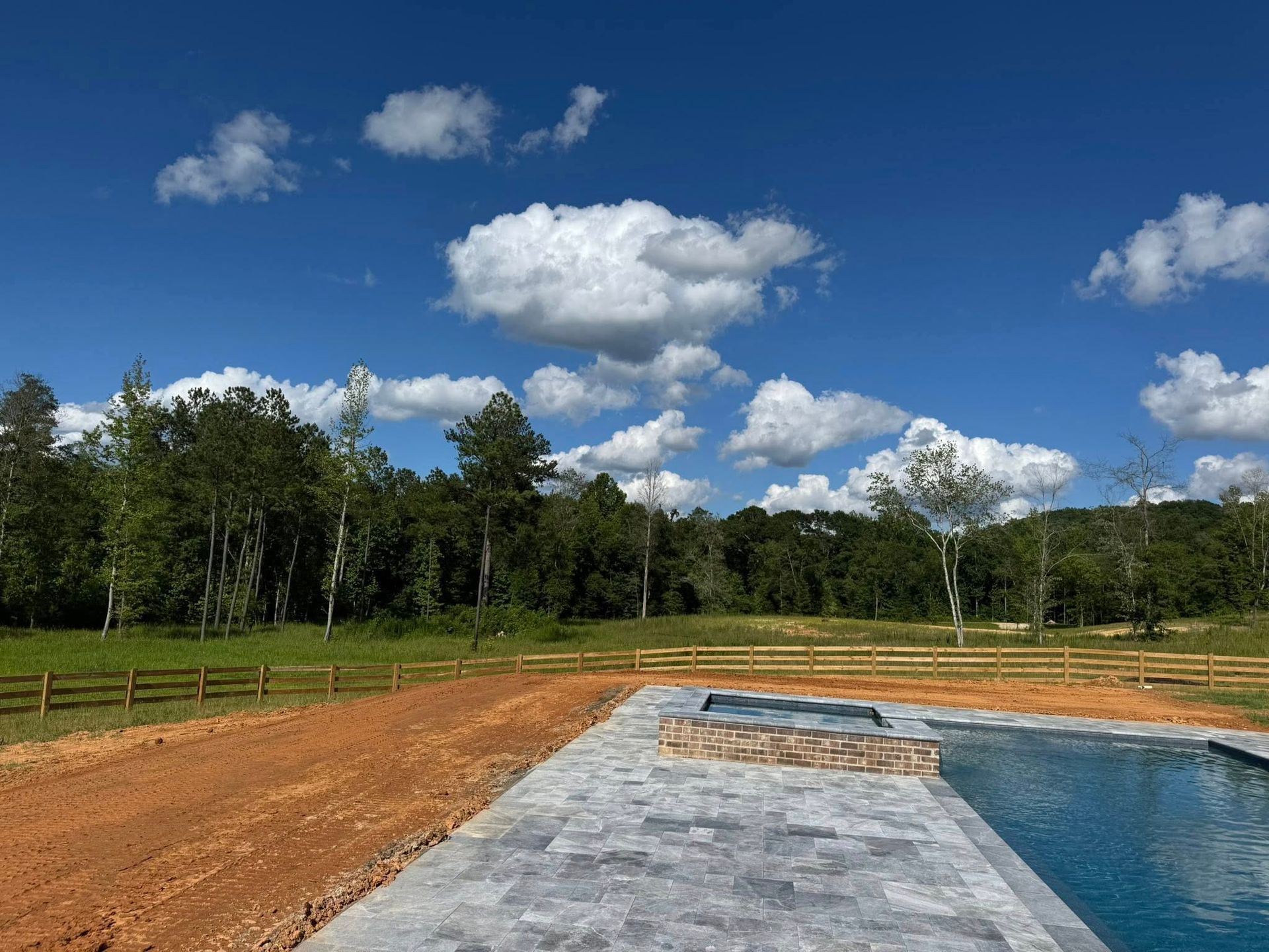 Backyard with a pool, stone patio, wooden fence, and trees under a blue sky with white clouds.