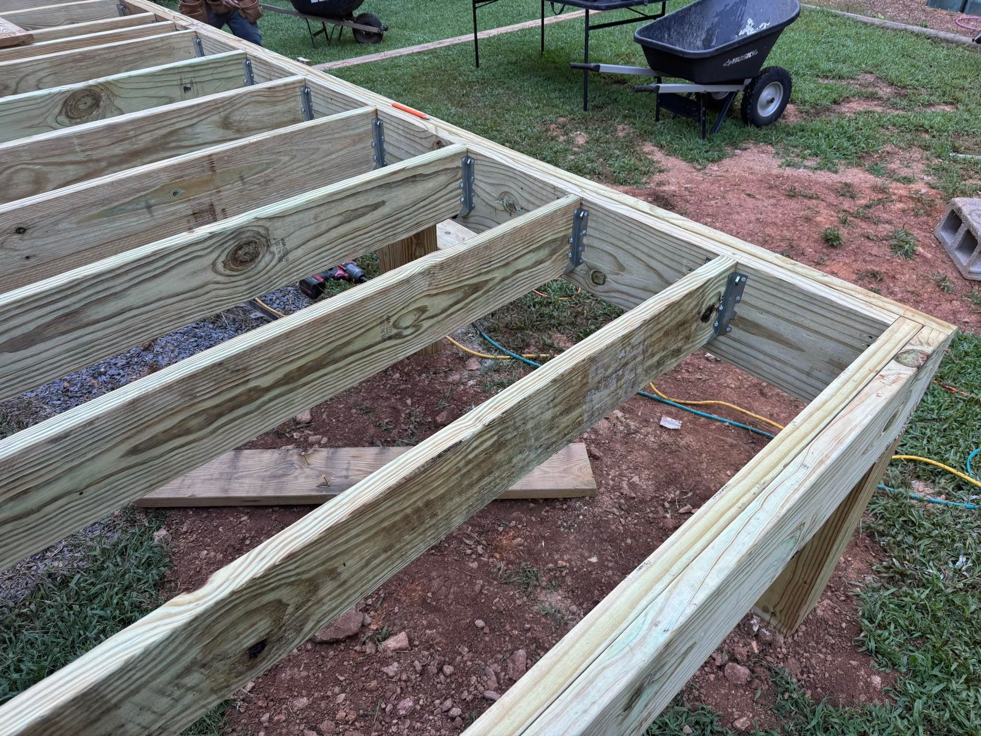 Wooden deck frame under construction outdoors on dirt and grass.