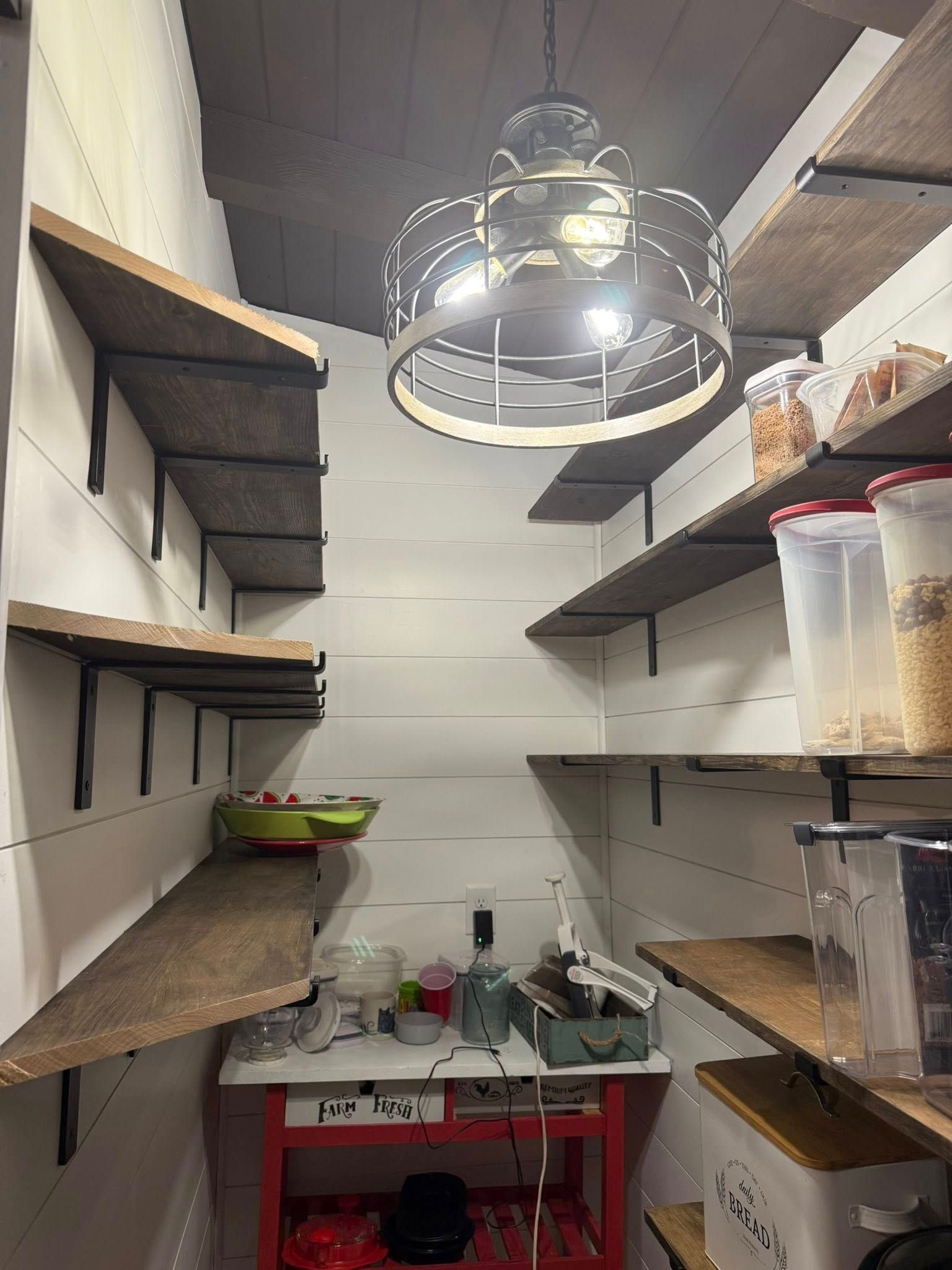 Pantry with open wooden shelves on white walls, illuminated by a hanging light. Some food containers are visible.