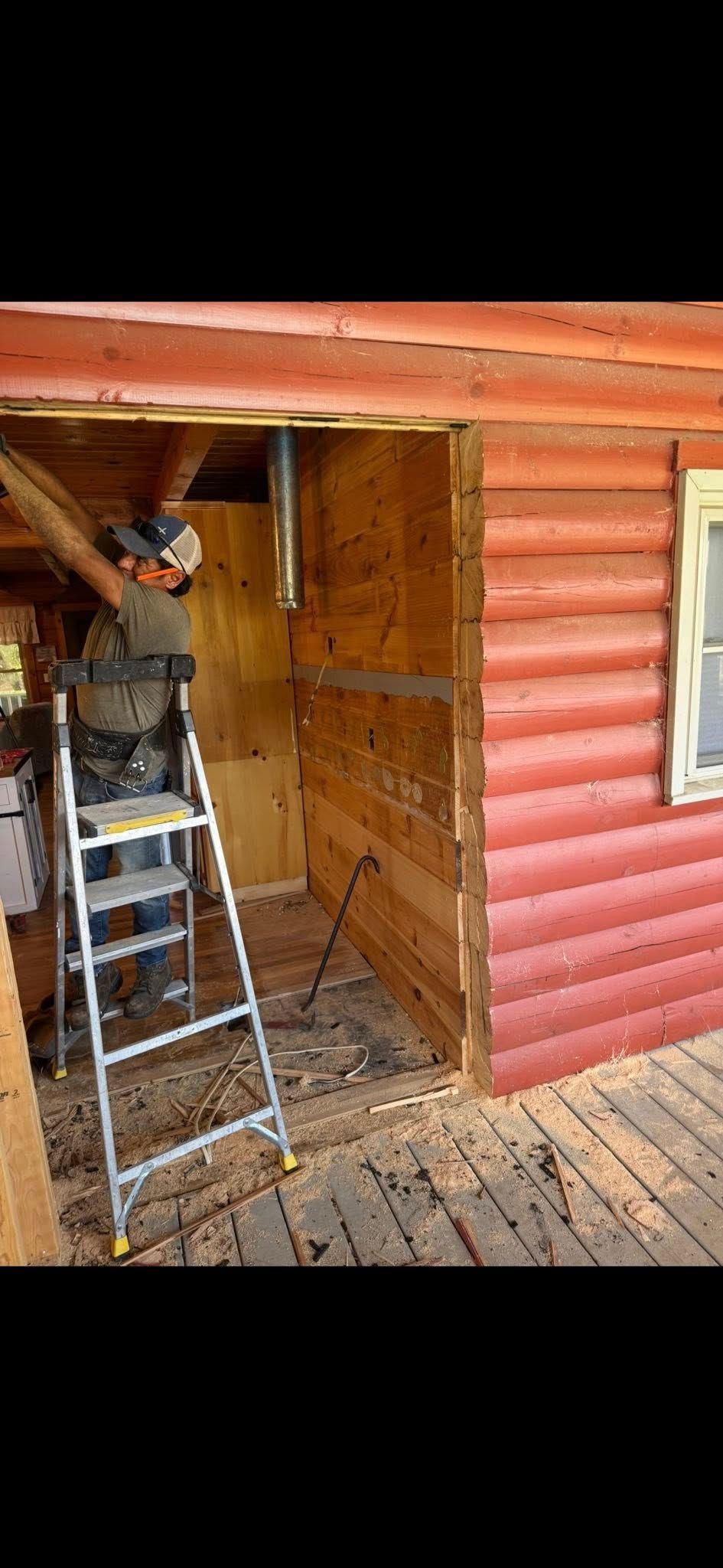 Person on ladder using power saw to cut into wooden wall of a red log cabin.