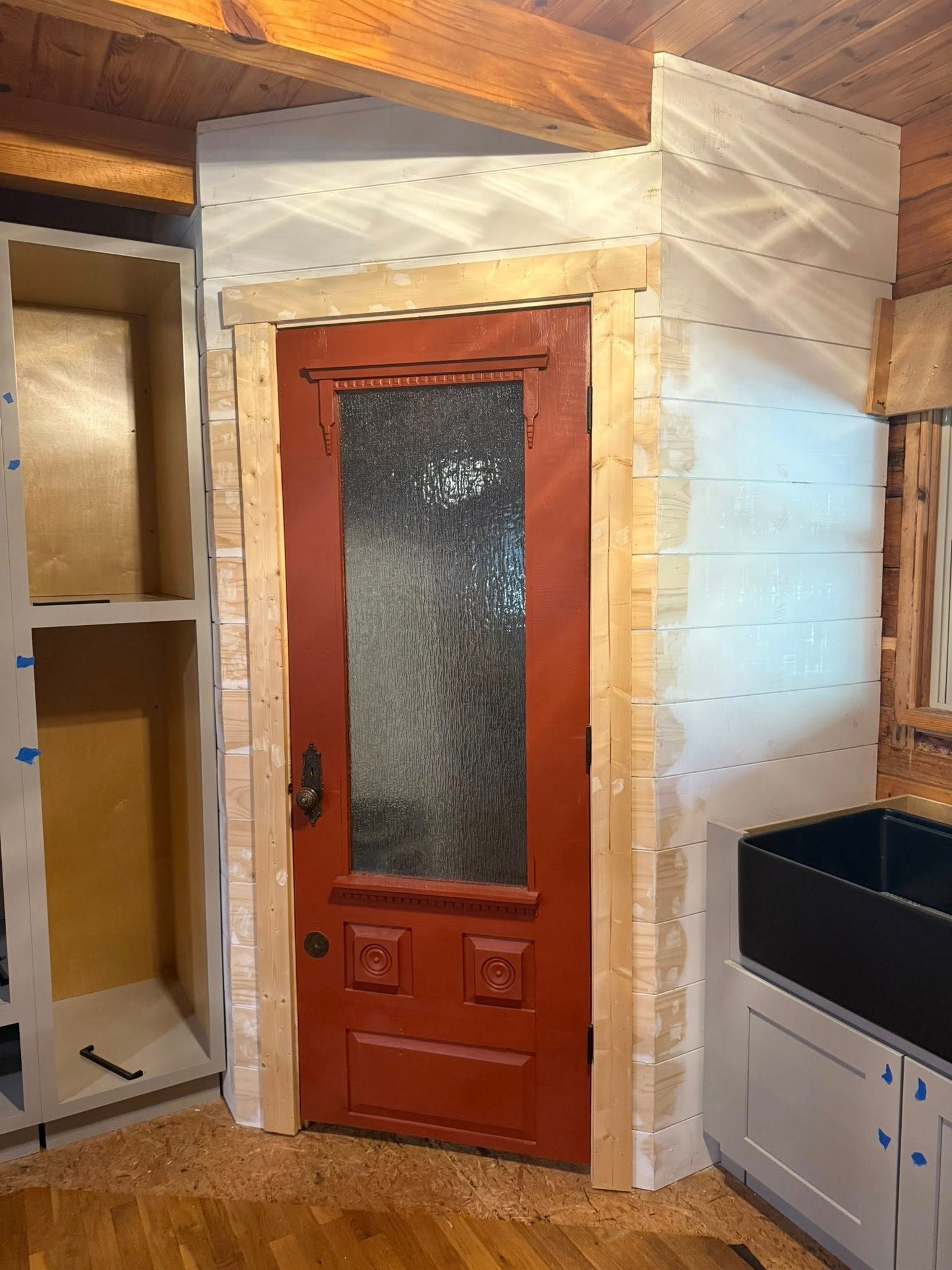 Red door with glass panel, in a kitchen renovation with wooden trim, cabinets, and a sink.