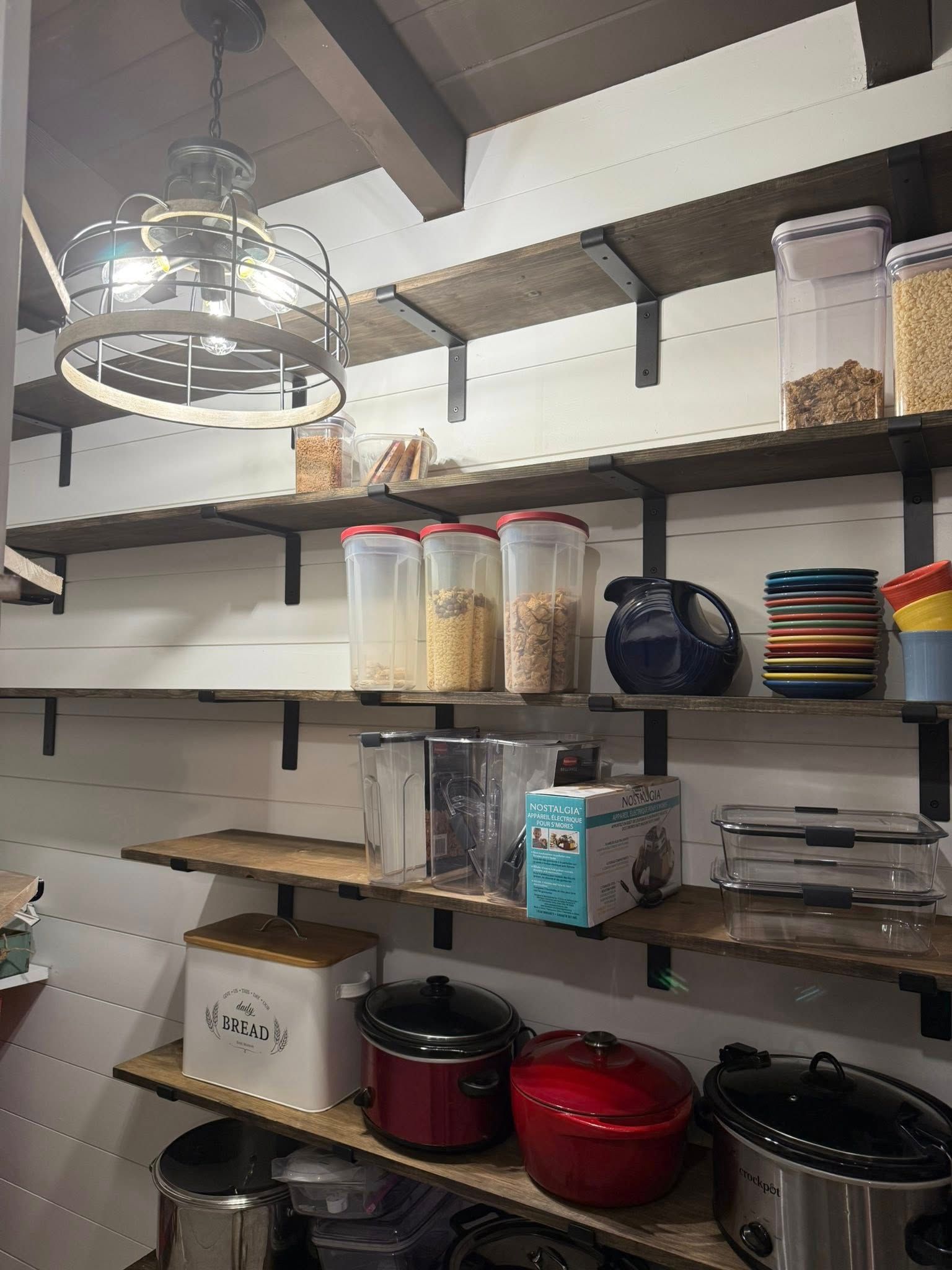 Pantry with open wooden shelves. Jars of food and appliances are stored on the shelves. A light fixture hangs from the ceiling.