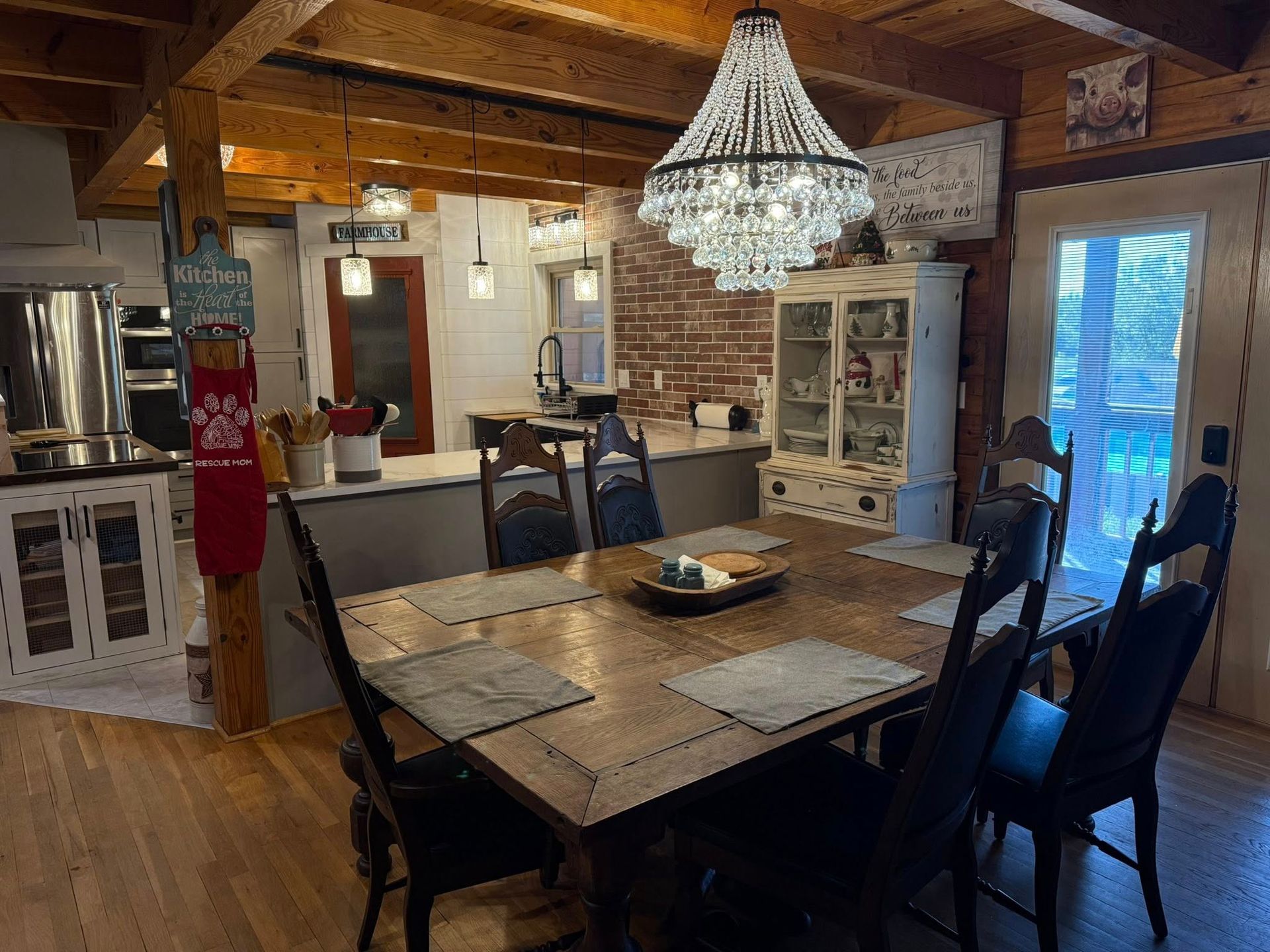 Dining room with wooden table, black chairs, chandelier, and a kitchen visible in the background.