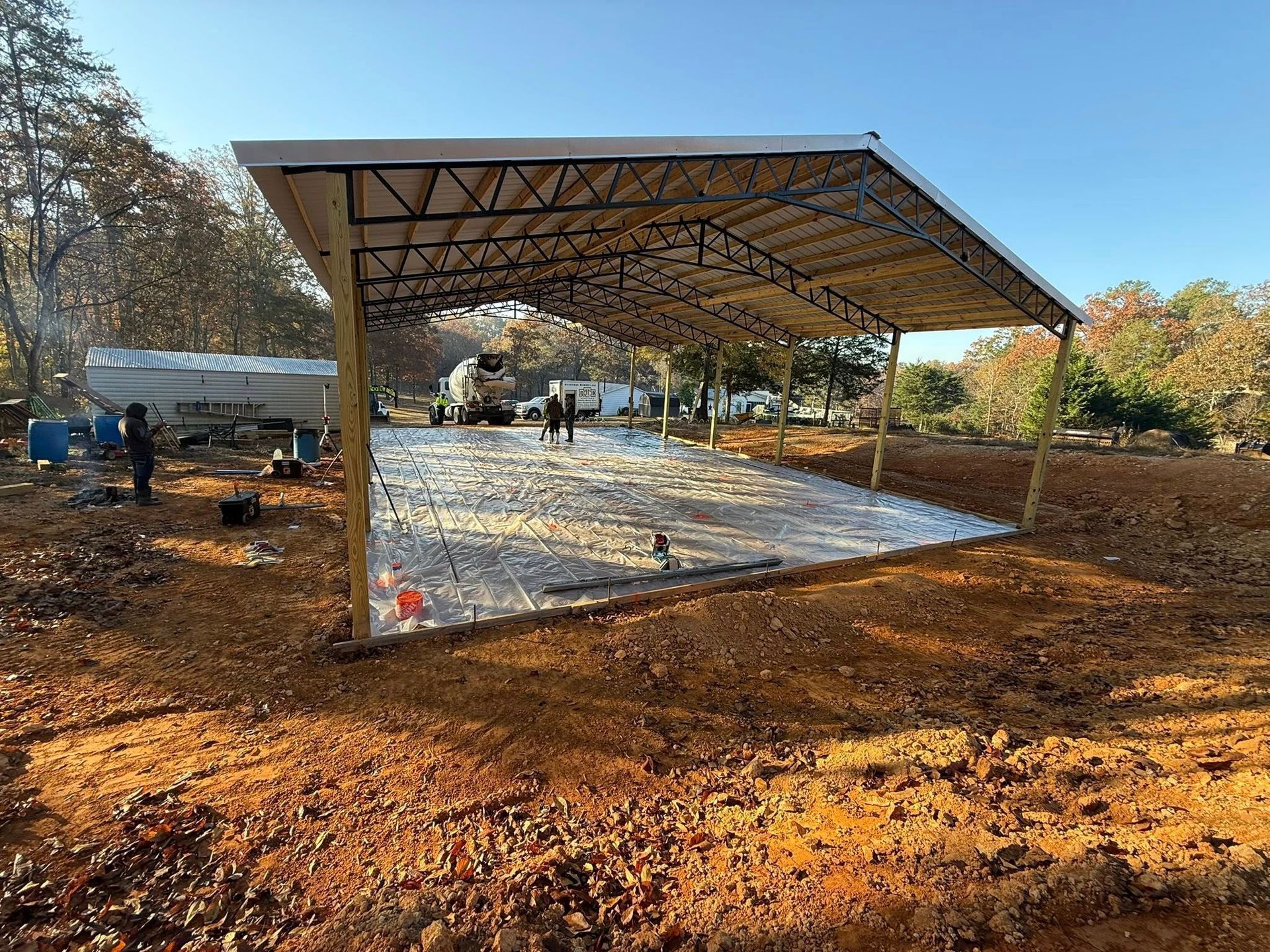 Construction site: Concrete slab under a metal roof structure. Workers visible in a rural setting.