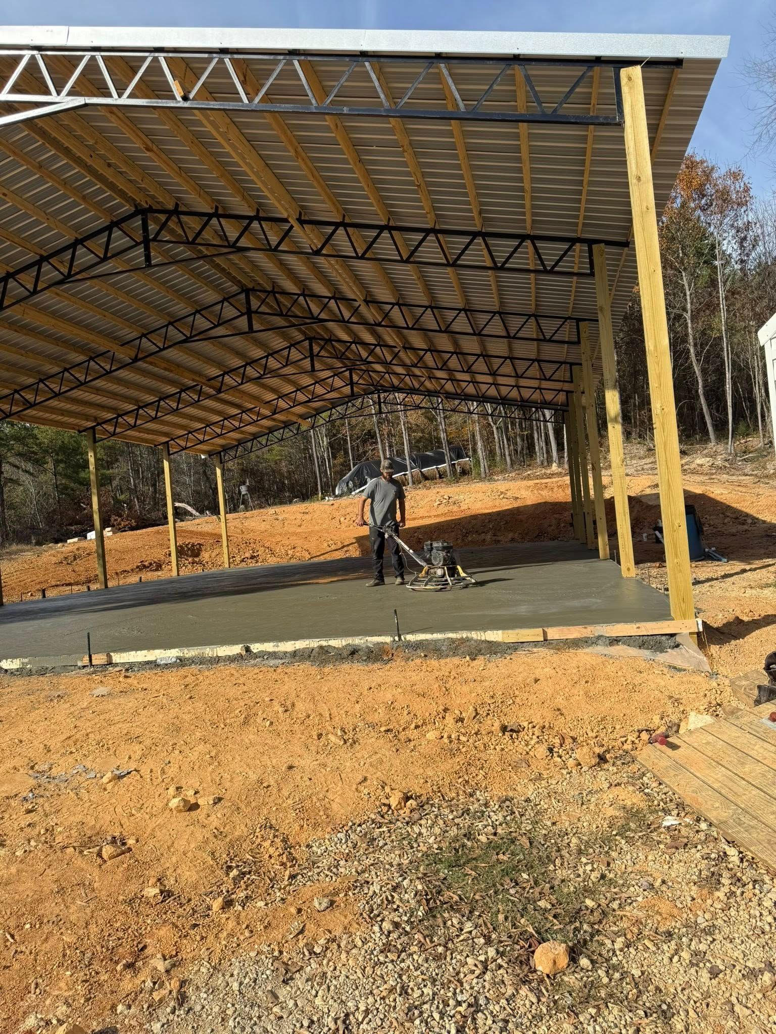 Man smoothing wet concrete under a metal-roofed shelter. Dirt surrounds the foundation with trees in the background.